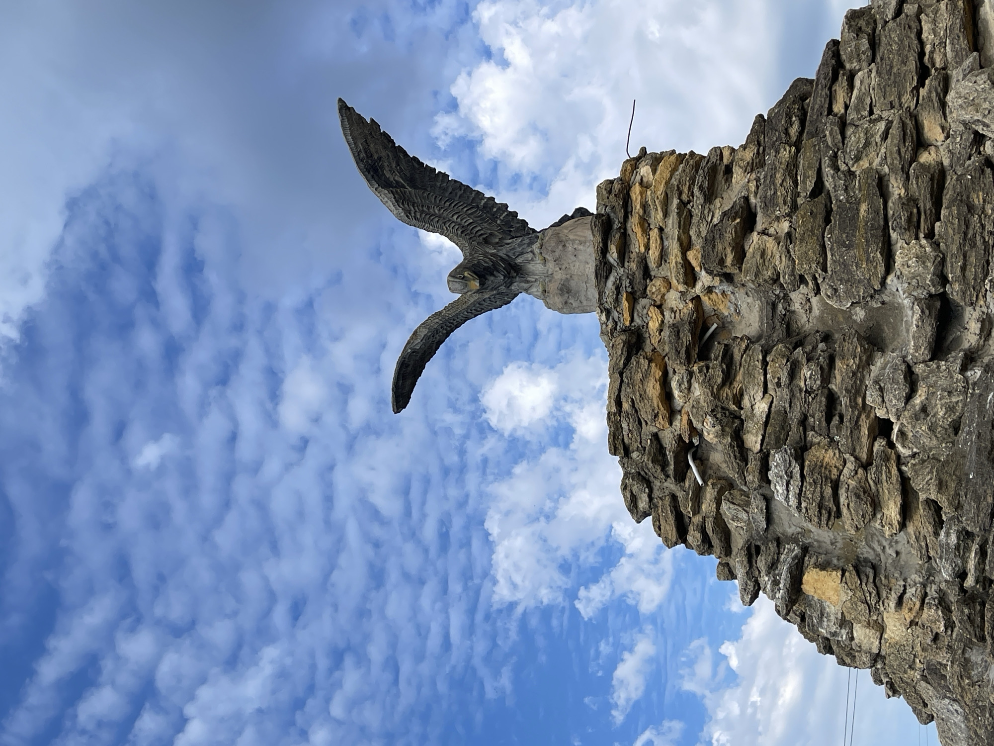 A low-angle, full-shot photograph captures a stone sculpture of an eagle with its wings outstretched, perched atop a rocky pedestal. The sculpture is a dark gray, weathered material, with detailed feathering visible on the wings and body. The eagle's head is slightly bowed, facing forward. The pedestal is constructed from rough, uneven stones of various sizes, predominantly in shades of brown and gray, with some lighter tan stones interspersed. The pedestal has a rough, asymmetrical shape, resembling a natural rock formation.
The background is dominated by a bright blue sky with scattered white, wispy clouds. The clouds are textured and form a dynamic pattern across the sky. In the far background, visible behind the lower left portion of the pedestal, are faint, thin dark lines that appear to be power lines.
The overall impression is one of a monument or landmark, possibly in an outdoor setting, given the naturalistic elements of the sculpture and its surroundings. The sky suggests a clear, possibly daytime weather condition. This sculpture is located in the city of Donskoye, Russia.