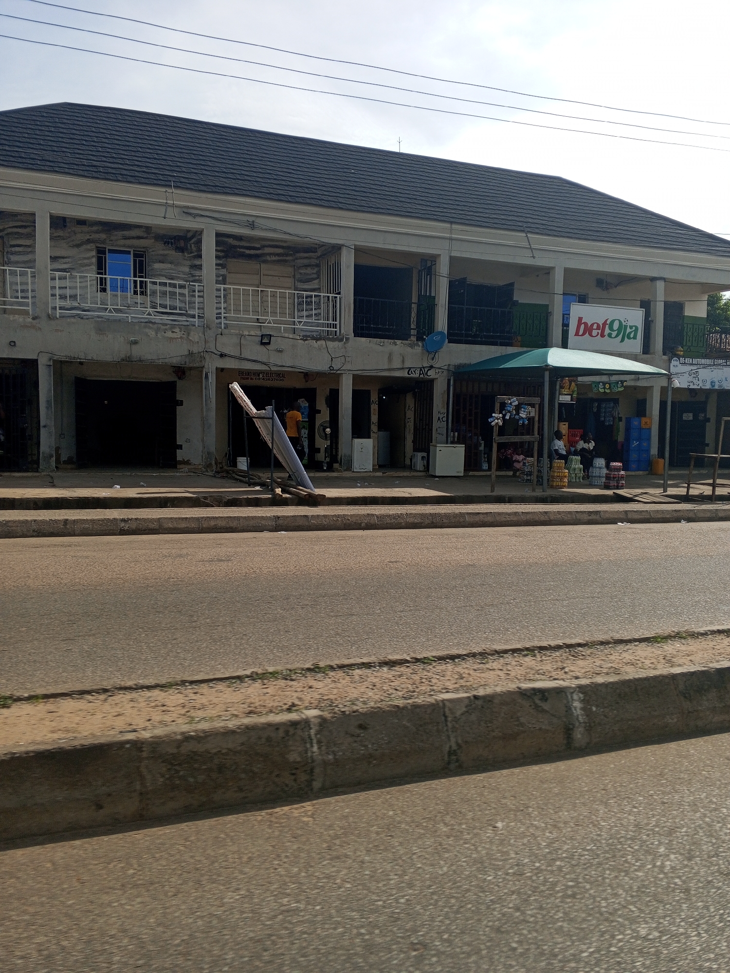 The image depicts a street scene in Jalingo, Nigeria, focusing on the facade of a two-story building with multiple storefronts. The building has a dark, sloped roof and a concrete exterior. On the ground floor, several businesses are visible. One storefront appears to be an electronics shop, with a sign for "EGEMMO NI." Another business, labeled "bet9ja," is a betting shop with a prominent red and white sign. Next to it, a shaded kiosk is set up, selling drinks, with stacks of colorful crates and bottles visible. Two individuals are seated under the kiosk. In front of the building, a sidewalk runs alongside the paved road. A man in an orange shirt is standing near a collapsed signpost. The sky is overcast.
