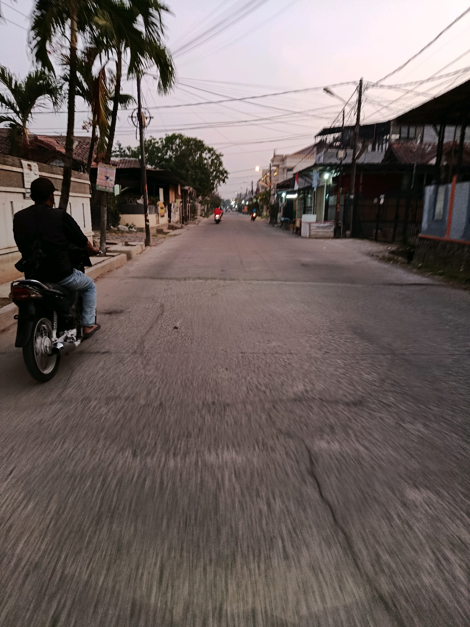 The image depicts a street scene in Bekasi, Indonesia, during dusk or early morning twilight. The perspective is from behind a person riding a motorcycle, moving forward on an asphalt road. The road is lined with buildings on both sides, some appearing to be shops or residences. Palm trees are visible on the left side of the street, with numerous electrical wires crisscrossing overhead. The lighting suggests a low-light environment, with the sky displaying a gradient of pale orange and grey. The motion blur in the foreground indicates movement, likely from the motorcycle's travel. A second motorcycle with a rider in a red shirt is visible further down the street, also moving in the same direction. The overall atmosphere is that of a typical urban street in Indonesia during the transitional light of dawn or dusk.