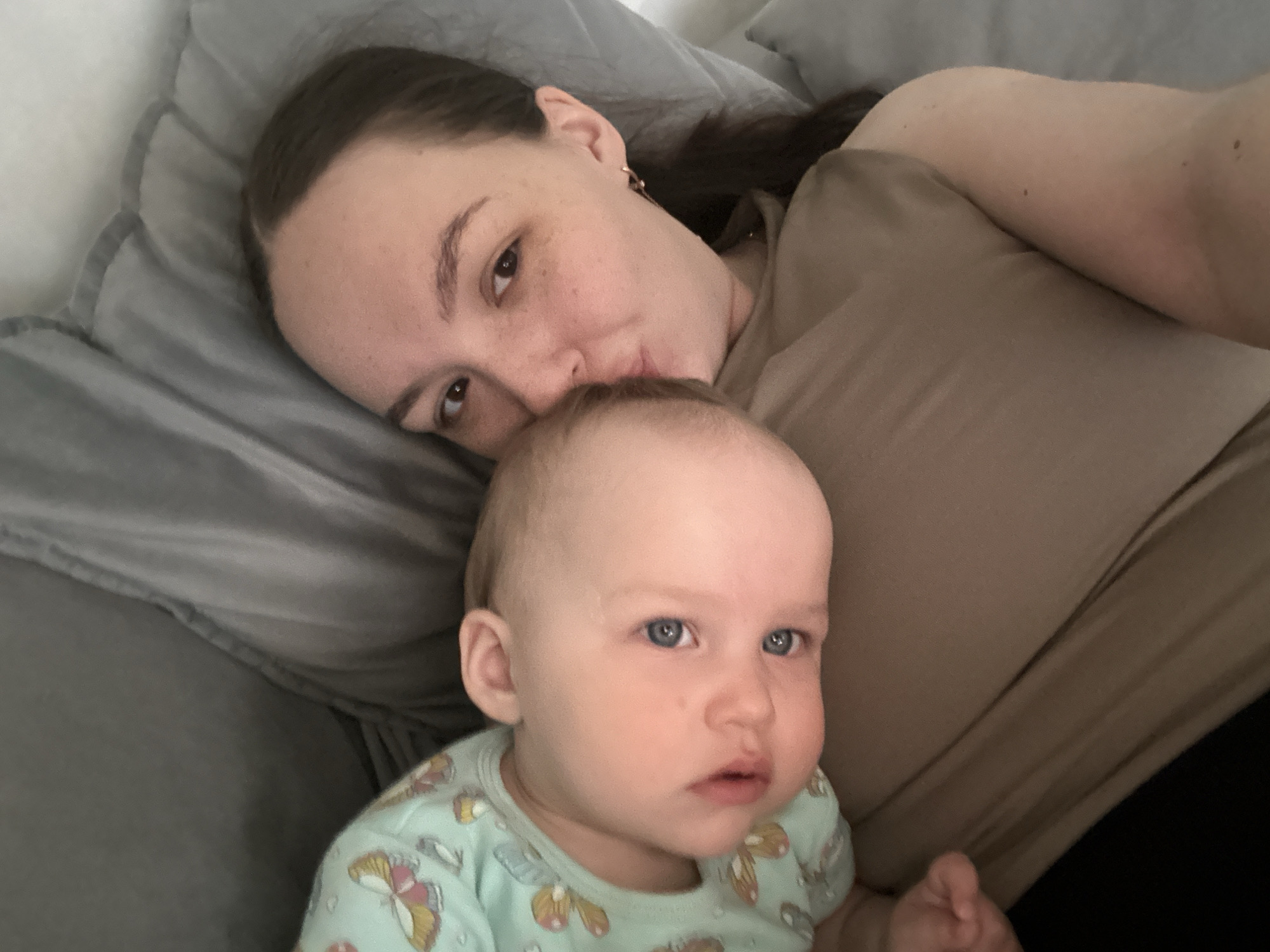 A woman and a baby lie together. The woman has brown hair and is wearing a brown shirt. She is kissing the baby on the head. The baby has blue eyes and is wearing a green shirt with butterfly pattern. They are on a gray pillow or bed. Location: Unknown, but potentially Chaikovsky, Russia.