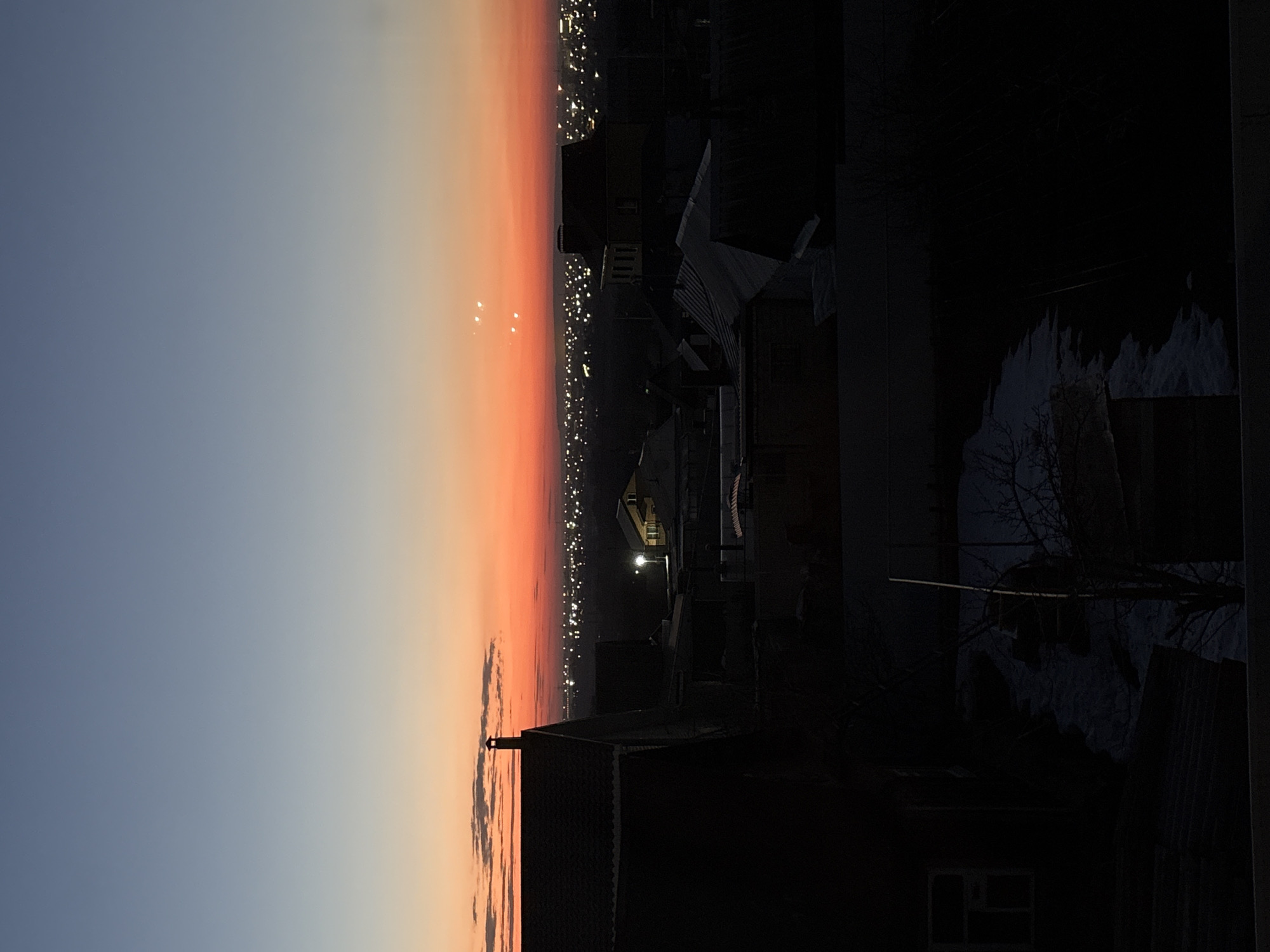 The image depicts a cityscape at dawn or dusk, likely in Ulyanovsk, Russia, judging by the urban lighting pattern visible along the horizon. A gradient sky stretches from blue to orange, indicating a sunrise or sunset. In the foreground, a dark residential neighborhood is present, featuring rooftops of houses. The overall impression is an urban scene transitioning from darkness to light.