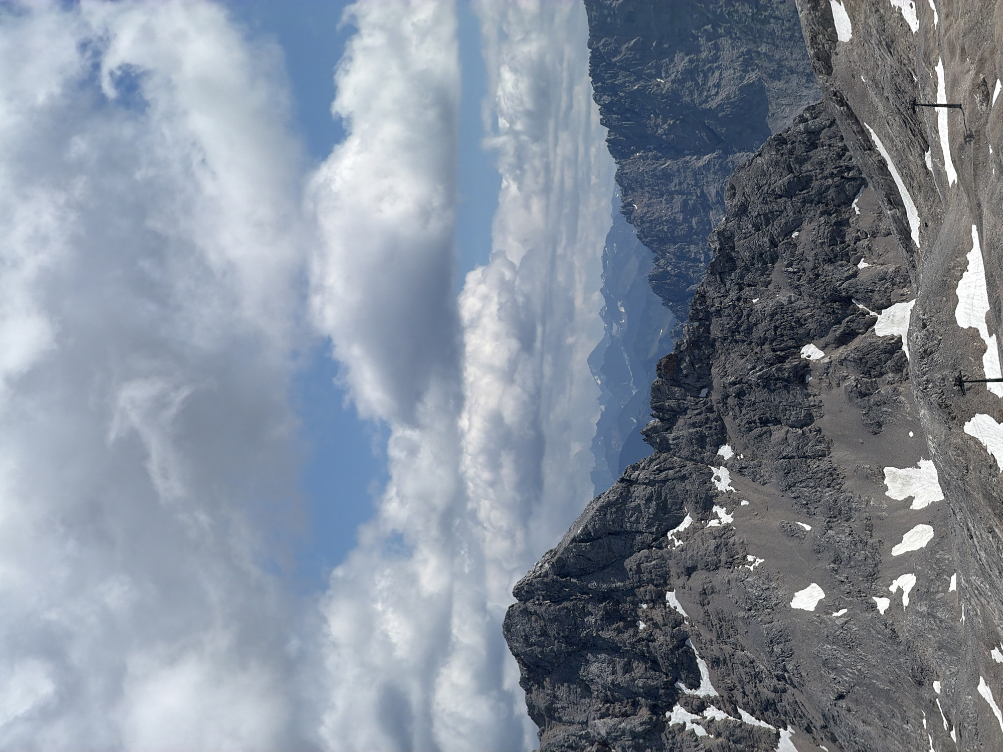 This is a photograph of a mountain landscape, likely taken from a high vantage point. The foreground features rugged, grey rock formations with patches of snow scattered across the slopes. In the middle ground, a series of jagged mountain peaks extend into the distance, with layers of mountains fading into a hazy blue. The sky above is a mix of bright blue and large, fluffy white clouds. There are two dark, slender poles visible in the lower right portion of the image, which appear to be part of a ski lift or similar infrastructure. The scene is characteristic of the Bavarian Alps, and specifically the area surrounding Garmisch-Partenkirchen, Germany, known for its high mountain peaks and ski resorts. There are no people or animals visible in the image.