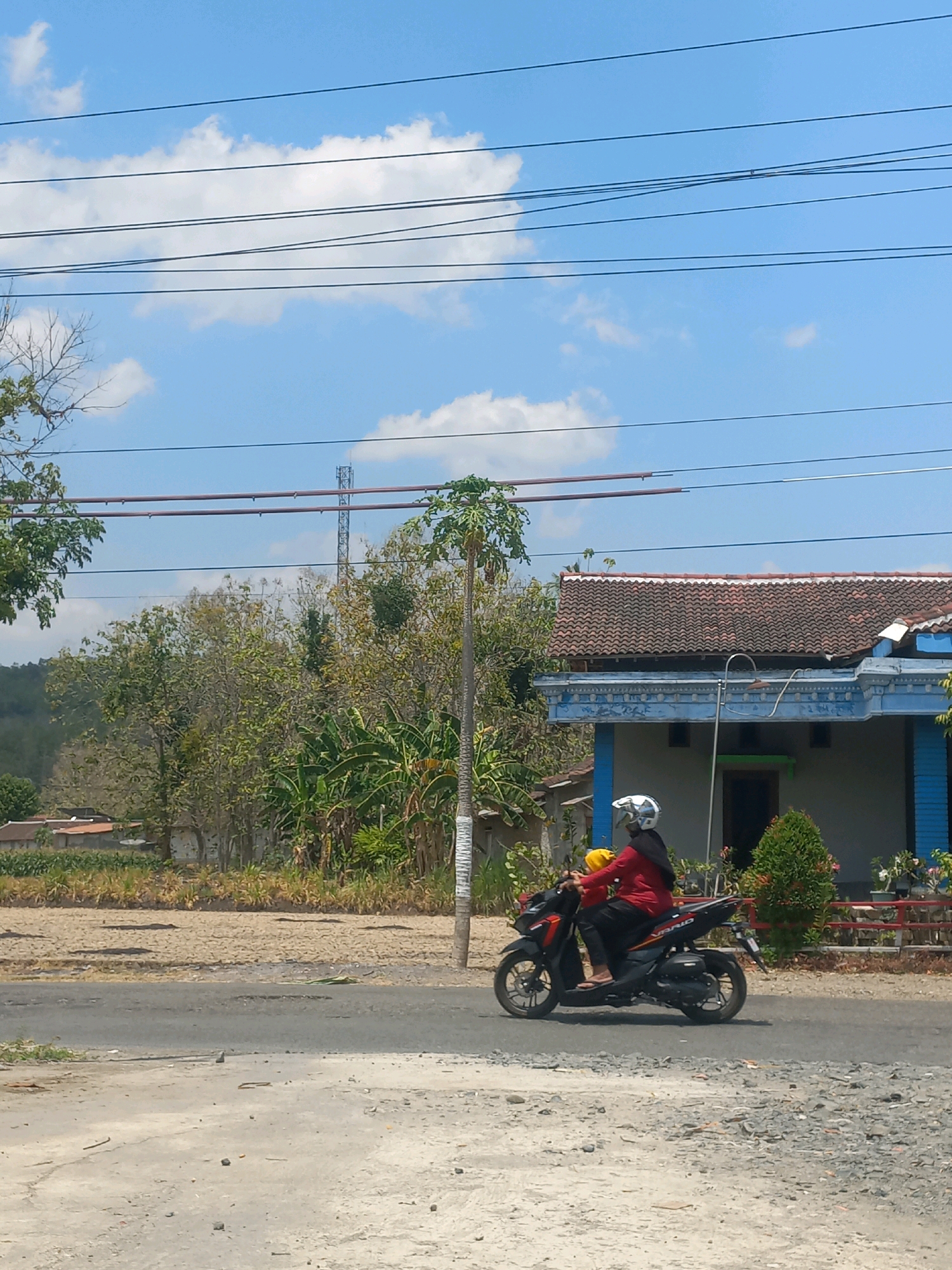 An outdoor, eye-level, full-shot image captures a rural scene in Baturetno, Indonesia, under a bright blue sky dotted with white clouds. The foreground features a weathered, asphalt road, partially covered with small pebbles and debris, with a rougher, gravelly surface to the left. In the mid-ground, a black motorcycle with orange and white accents is ridden by two individuals. The driver, wearing a red shirt, black pants, and a white helmet with a black hijab, is holding onto the handlebars. The passenger, wearing a yellow shirt and a black hijab, sits behind the driver. The motorcycle is in motion, traveling from left to right on the asphalt road. To the right of the road is a residential building with a brown tiled roof and light blue trim. Green bushes and small trees surround the house. Beyond the house, a field of dry, brown earth is visible, with some vegetation and a few houses with orange roofs in the distance. A cell tower stands tall in the background amidst a cluster of trees. A single tree with sparse foliage stands to the left of the motorcycle. Power lines crisscross the sky horizontally.