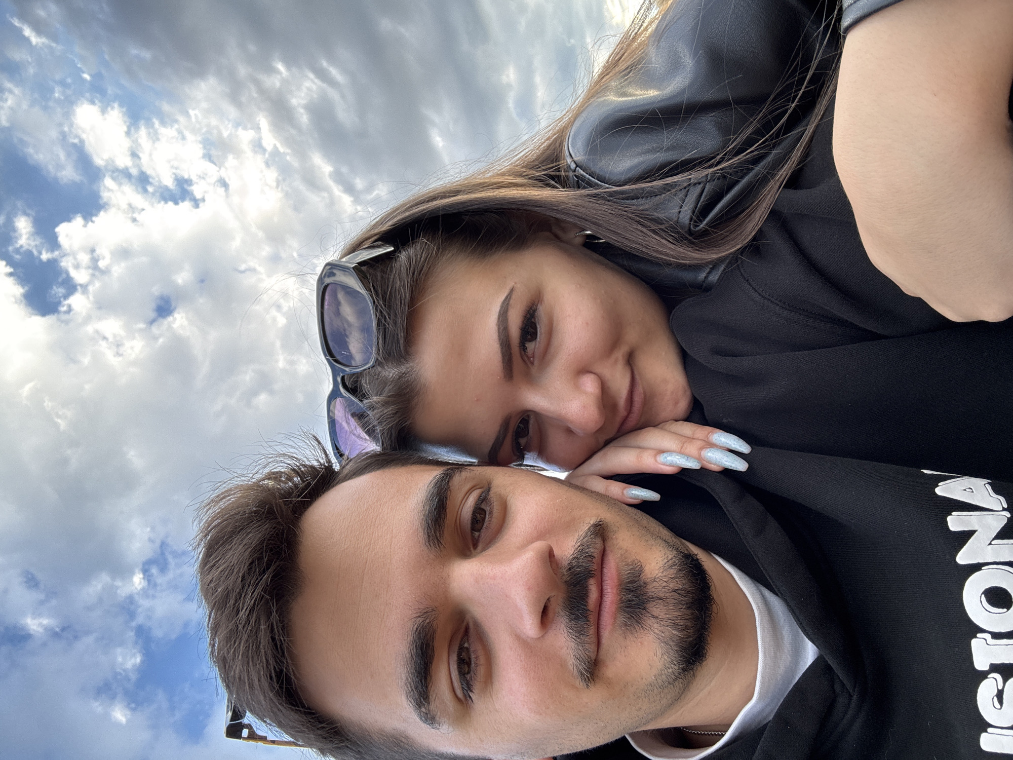 The image is a close-up selfie taken outdoors, with a cloudy sky as the background. The subjects are a young couple. The male subject is in the foreground, looking directly at the camera. He has dark hair, a mustache, and a goatee. He is wearing a black hoodie with a white logo that is partially visible. The female subject is to his right, leaning her head on his shoulder and looking towards the camera. She has long, dark hair and is wearing sunglasses on top of her head and a black leather jacket. Her left hand rests on the male subject's chest, displaying long, light blue glitter nails. The lighting suggests it is either early morning or late afternoon. The setting does not provide specific visual cues to pinpoint a location in Sofia, Bulgaria, beyond being an outdoor urban or semi-urban environment.