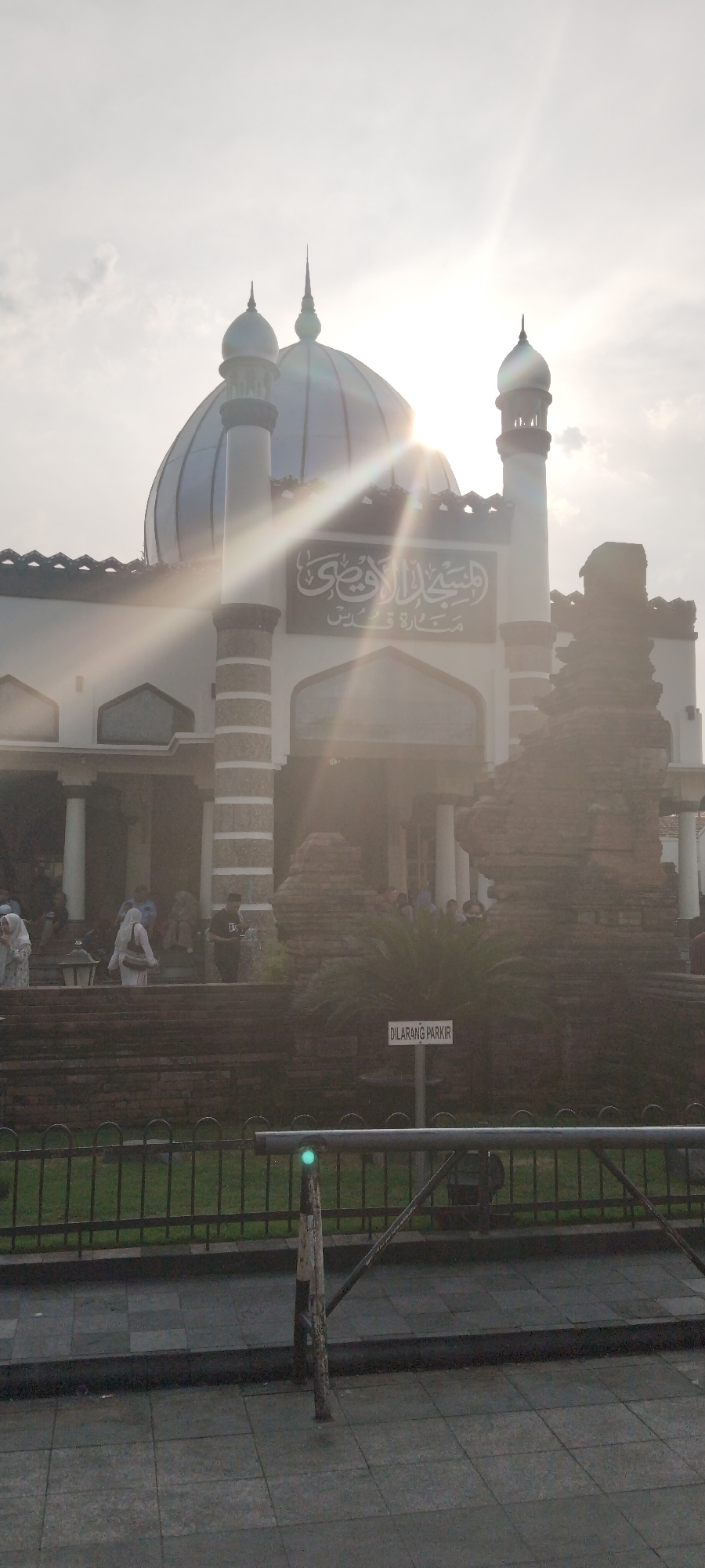 The image shows the exterior of the Great Mosque of Kauman in Yogyakarta, Indonesia, bathed in the bright sunlight of late afternoon, which creates lens flares and overexposure in parts of the image. The mosque features a prominent blue dome and minarets, with intricate architectural details. Arabic calligraphy is visible on the facade above the entrance.
In the foreground, there is a metal fence and a paved walkway. To the right, a weathered brick structure stands, possibly an older part of the mosque complex or a historical artifact. Several people, some wearing headscarves, are seen walking on the stairs leading up to the mosque and in the background. A sign that reads "DILARANG PARKIR" (which translates to "NO PARKING" in Indonesian) is visible on a post near the fence. The sky is bright with some clouds, and the sun is positioned high and slightly to the left of the frame. The overall scene suggests a public space during a time of religious observance or gathering.