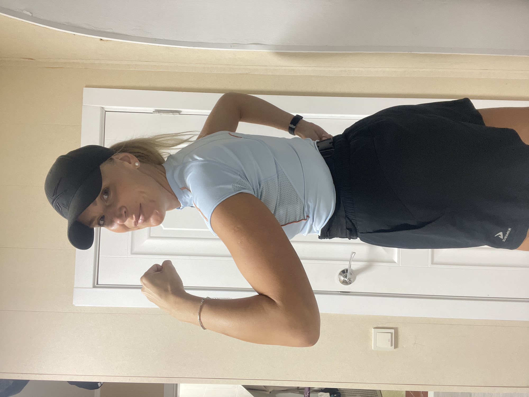 A woman is posing indoors, facing the camera, with a closed fist raised. She wears a black baseball cap, a light blue short-sleeved shirt, black shorts, and a black smartwatch on her left wrist. She has blond hair pulled back. The background includes a white door with a silver handle, a beige wall, and a white ceiling. A light switch is visible on the wall. No evidence indicates a specific location in Baranovka, Russia.