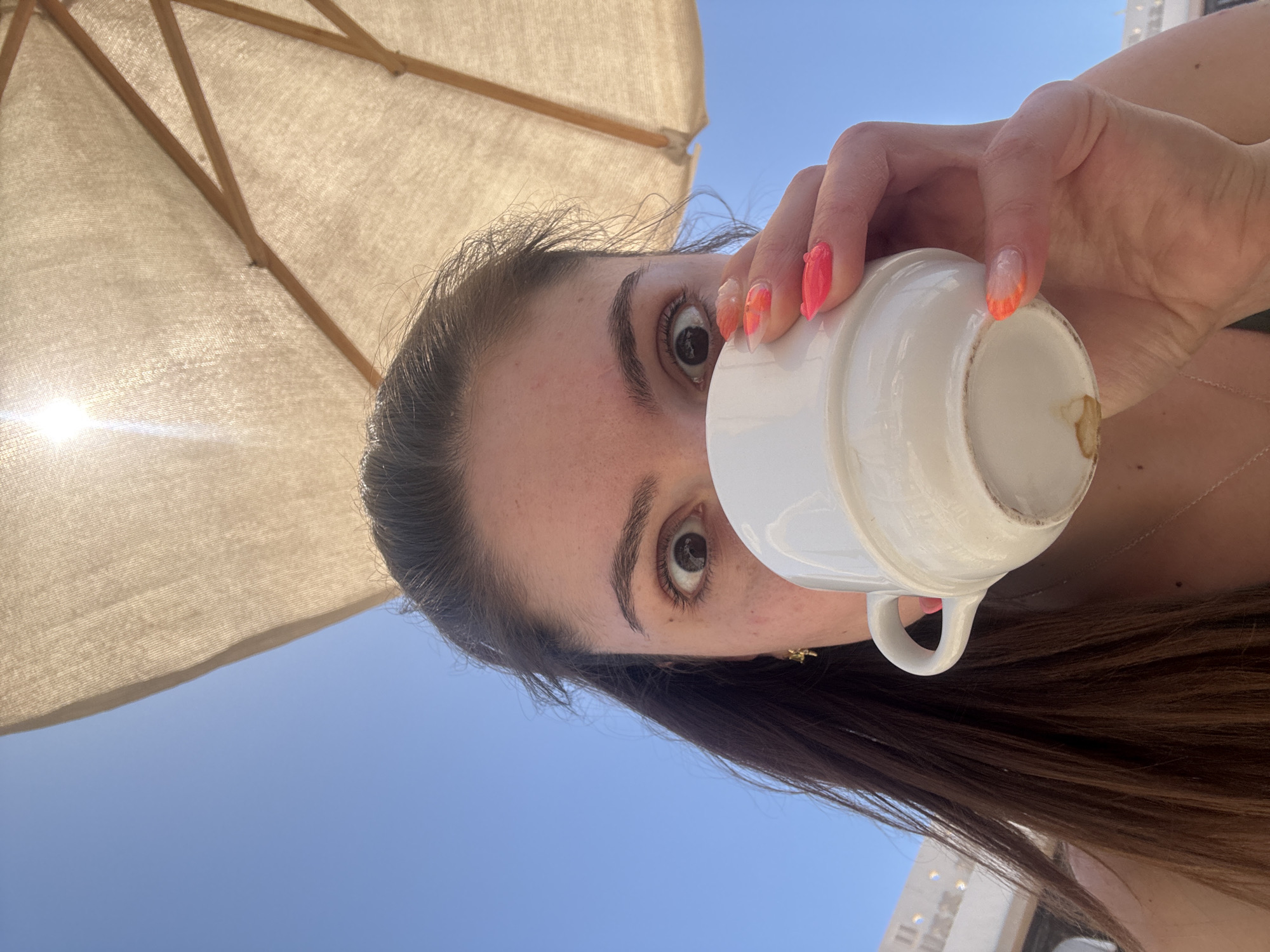 A woman is holding a white cup to her mouth, presumably drinking from it. She is positioned under a tan umbrella, with a clear blue sky visible in the background. Her nails are painted with an orange ombre design. Her hair is dark brown and slightly disheveled. Her expression is one of mild surprise or alertness. She appears to be in a city environment.