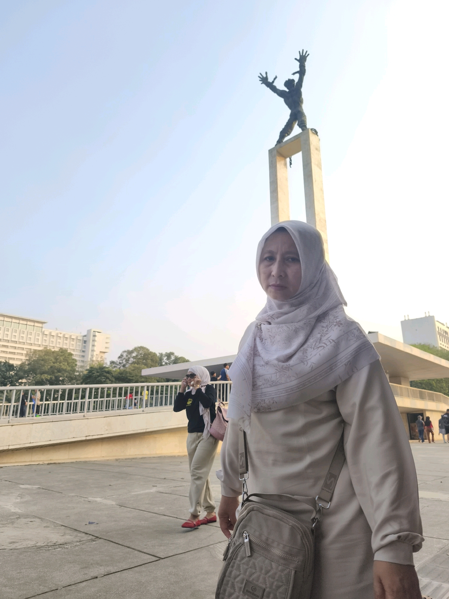 A medium shot captured outdoors shows a woman in the foreground wearing a light beige hijab and a matching long-sleeved top, with a light brown crossbody bag slung over her shoulder. She is looking directly at the camera with a neutral expression. To her left, further in the background, another woman wearing a white hijab and a black top is seen in profile, holding up her phone as if taking a photo.
The background features a tall, concrete monument topped with a bronze statue of a man with arms outstretched. Beyond the monument, a modern building with numerous windows and a pedestrian walkway with a white railing are visible. The setting appears to be a public square or park, with paved ground and a clear sky. The overall scene suggests a tourist or recreational location within Central Jakarta, Indonesia, possibly Monas National Monument Park, given the iconic statue and surrounding structures often found in such areas.