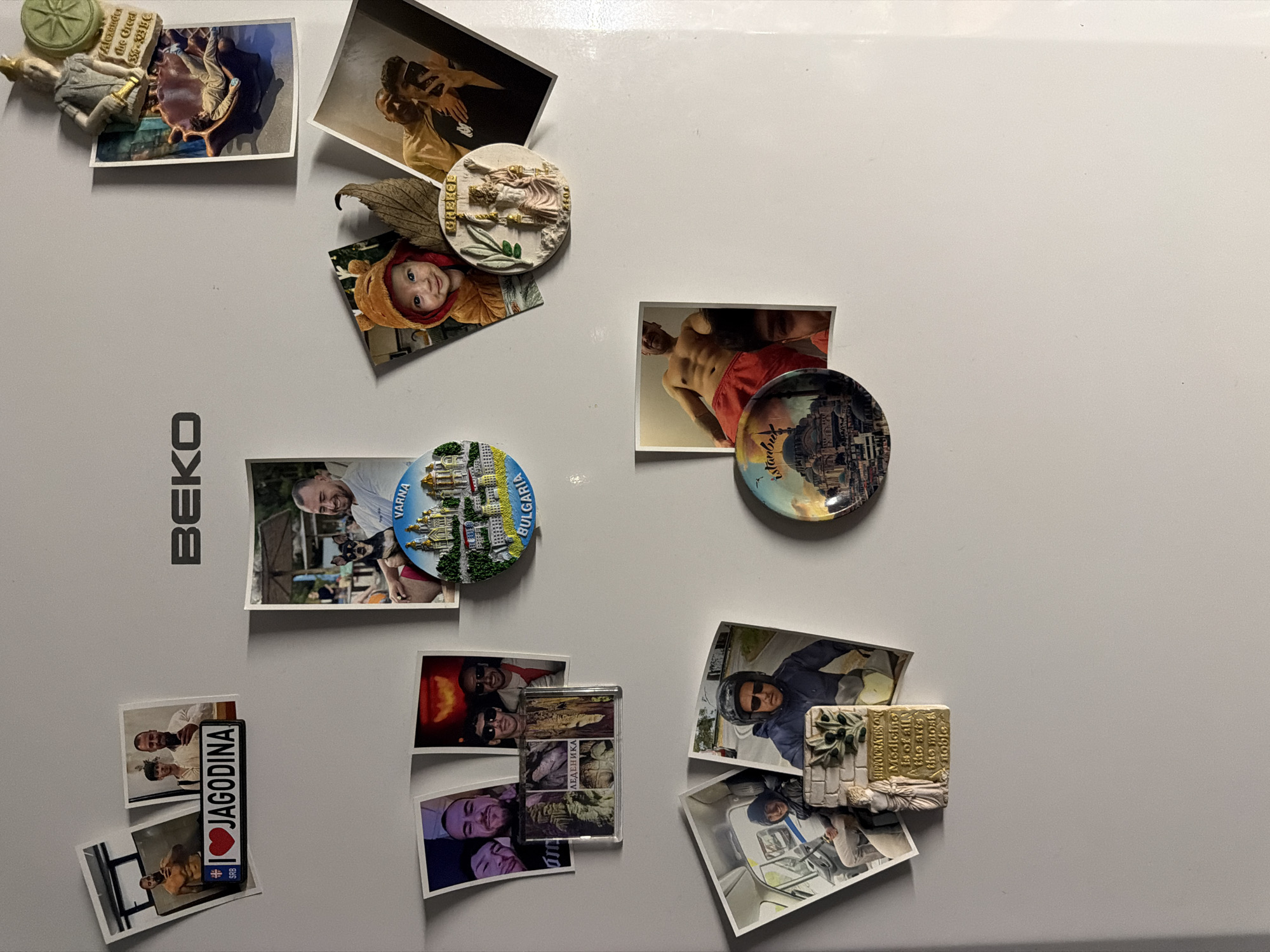 This is a top-down, slightly angled, full shot of a white refrigerator covered in magnets and photos. The refrigerator has a black BEKO logo in the upper center.
Magnets and photos are scattered across the surface. In the upper left, there are three photos, one with the text "I❤️JAGODINA". Below that, a group of photos includes three men in one, and two men in another. A clear magnet with an image of a cave and text below it is also present. In the upper right, a magnetic figurine of a person holding a book and a green wheel is placed above a photo of a person lying down. To the right of that, a photo captures two men taking a selfie.
In the center of the refrigerator, a round magnet with a city scene and the text "VARNA BULGARIA" is visible. To its left, a photo shows a man and a dog, with a beach umbrella in the background. Below the baby photo, there's a circular magnet with a statue and "GREECE" text.
In the lower portion of the refrigerator, a photo shows a man with a bare torso. Next to it, a round magnet depicts a mosque and the text "Istanbul". Below that, a photo shows a man on a motorcycle. To the left, a photo shows two people in a vehicle. Finally, a rectangular magnet with a statue and text that reads "HIPPOCRATES of Medicine is of all arts the most noble" is present.
The lighting suggests an indoor environment, likely a kitchen, with the flash from the camera creating reflections on the refrigerator's surface.