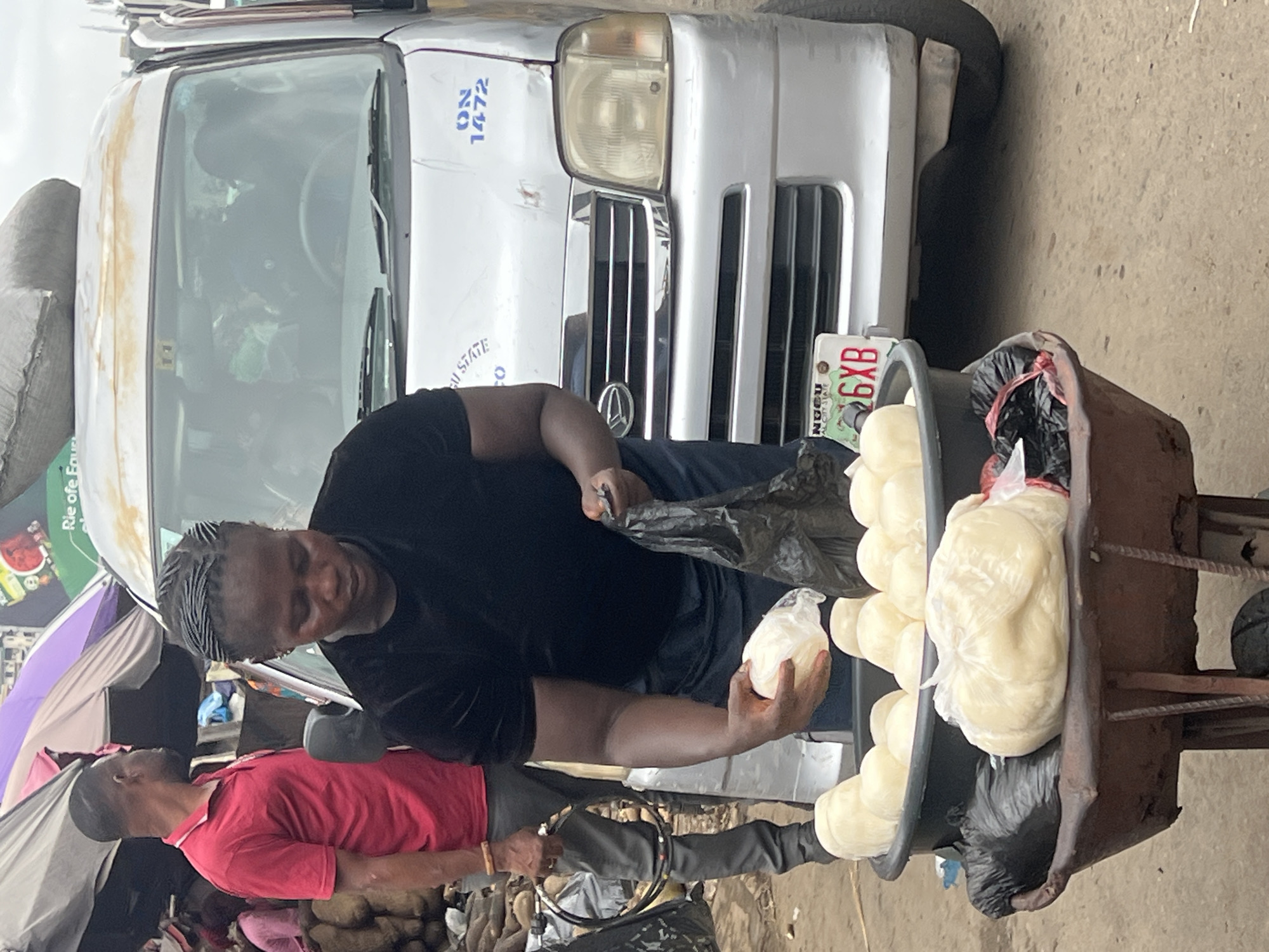 An outdoor market scene is depicted in Nkpor, Nigeria, with a focus on a woman selling food items from a cart. She is wearing a black t-shirt and dark pants and is in the process of handling a white food item wrapped in plastic. The cart contains several similar food items in a large black basin, along with some bagged items. Behind her is a silver van with blue lettering on the windshield and hood, indicating it's from "ENUGU STATE" with the number "ON 1472". A man in a red polo shirt and grey pants is standing to the left of the woman, looking away from the camera. Various stalls with umbrellas and produce are visible in the background, suggesting a busy market environment. The ground appears to be dirt or rough pavement.