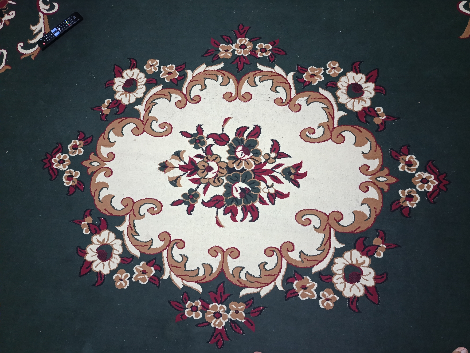 This is a top-down, eye-level, full shot of a dark green rug with a large, intricate floral pattern in the center. The pattern features an oval-shaped cream-colored area with a bouquet of flowers in dark red, beige, and dark green. The floral motif extends outward with swirling brown and cream-colored borders and additional smaller flowers in the same color palette scattered across the dark green background. A black remote control is placed in the upper left corner of the rug, angled diagonally. The lighting appears to be indoor, possibly from overhead sources, creating some subtle shadows. The image is forensic in nature, providing a detailed, objective view of the rug and the remote. The location within the city of Ӯртабуз, Tajikistan, is not ascertainable from the image itself.