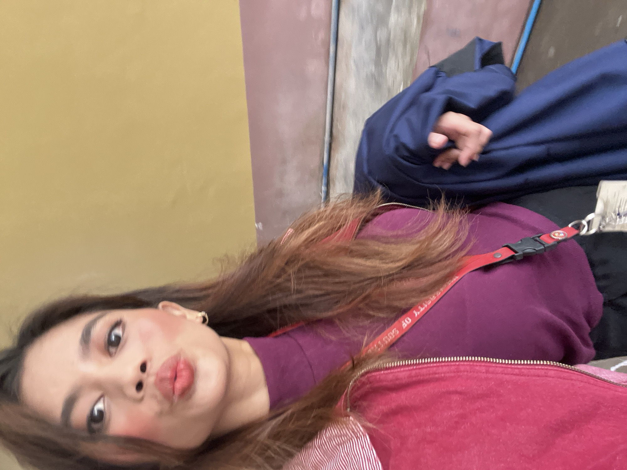A young woman is shown in a selfie with a pout. She has long brown hair and is wearing a purple sweater and red jacket with a red lanyard. In the background is a yellow wall and a surface with a blue jacket on it. The lanyard has text on it, but the exact wording is illegible. The location is Davao City, Philippines.