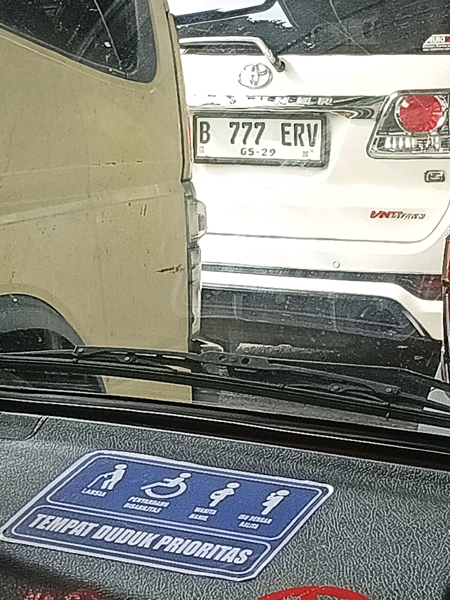The image shows a close-up view of the dashboard and windshield of a vehicle, with a sticker on the dashboard indicating "TEMPAT DUDUK PRIORITAS" (Priority Seating). The sticker features icons representing a disabled person in a wheelchair, an elderly person, and a pregnant woman. In the background, a white Toyota Fortuner with license plate "B 777 ERV" is visible, along with another vehicle to its left. The license plate suggests the vehicle is registered in Jakarta, Indonesia. The image is captured from inside a vehicle, likely parked in a traffic situation, possibly in South Jakarta, Indonesia, given the vehicle registration context.
