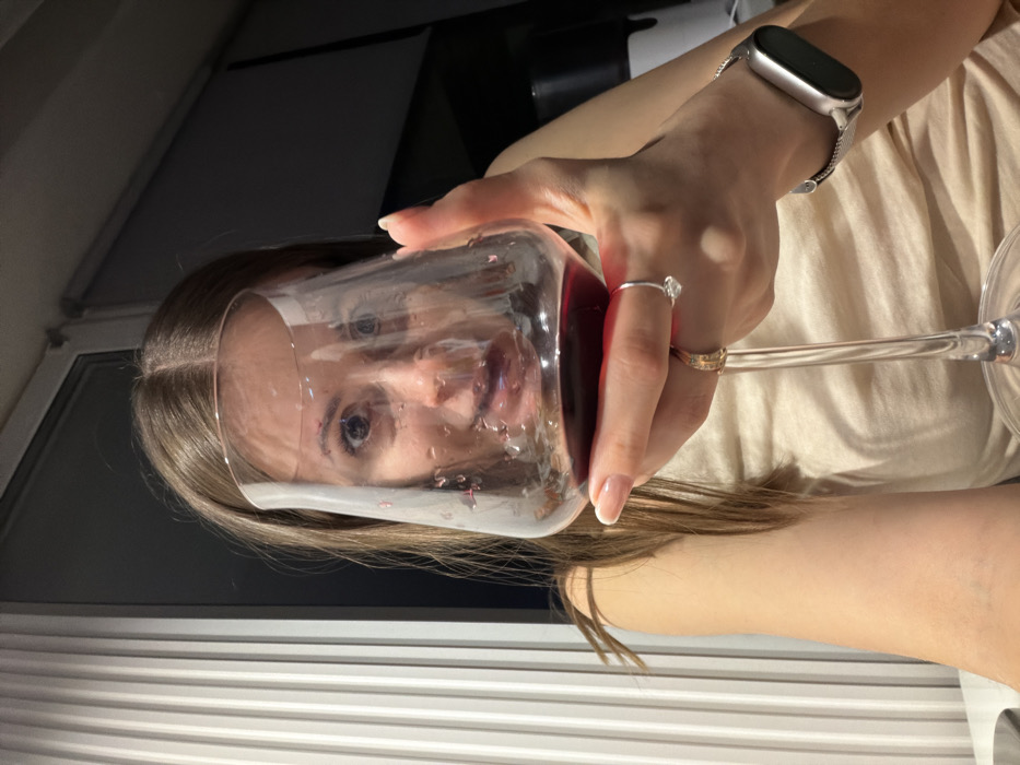 An indoor, eye-level photograph captures a young adult female in Chaikovsky, Russia, holding a stemmed wine glass filled with dark red liquid, presumably wine, directly in front of her face. Her head and upper torso are visible. She has long, straight, brown hair parted in the center. Her face is optically distorted by the glass and its contents, with her wide-open eyes looking directly at the viewer. Small droplets are visible on the inside surface of the glass above the wine level. Her left hand, with manicured nails featuring French tips, grips the stem and lower bowl of the glass. On her left ring finger, she wears two rings: an upper silver-colored ring with a clear stone, and a lower gold-colored band. A silver-colored smartwatch with a mesh band is visible on her left wrist. She wears a light-colored top. The background features vertical, light-colored window blinds on the left and a darker, indistinct area on the right, suggesting a low-light environment, possibly evening.