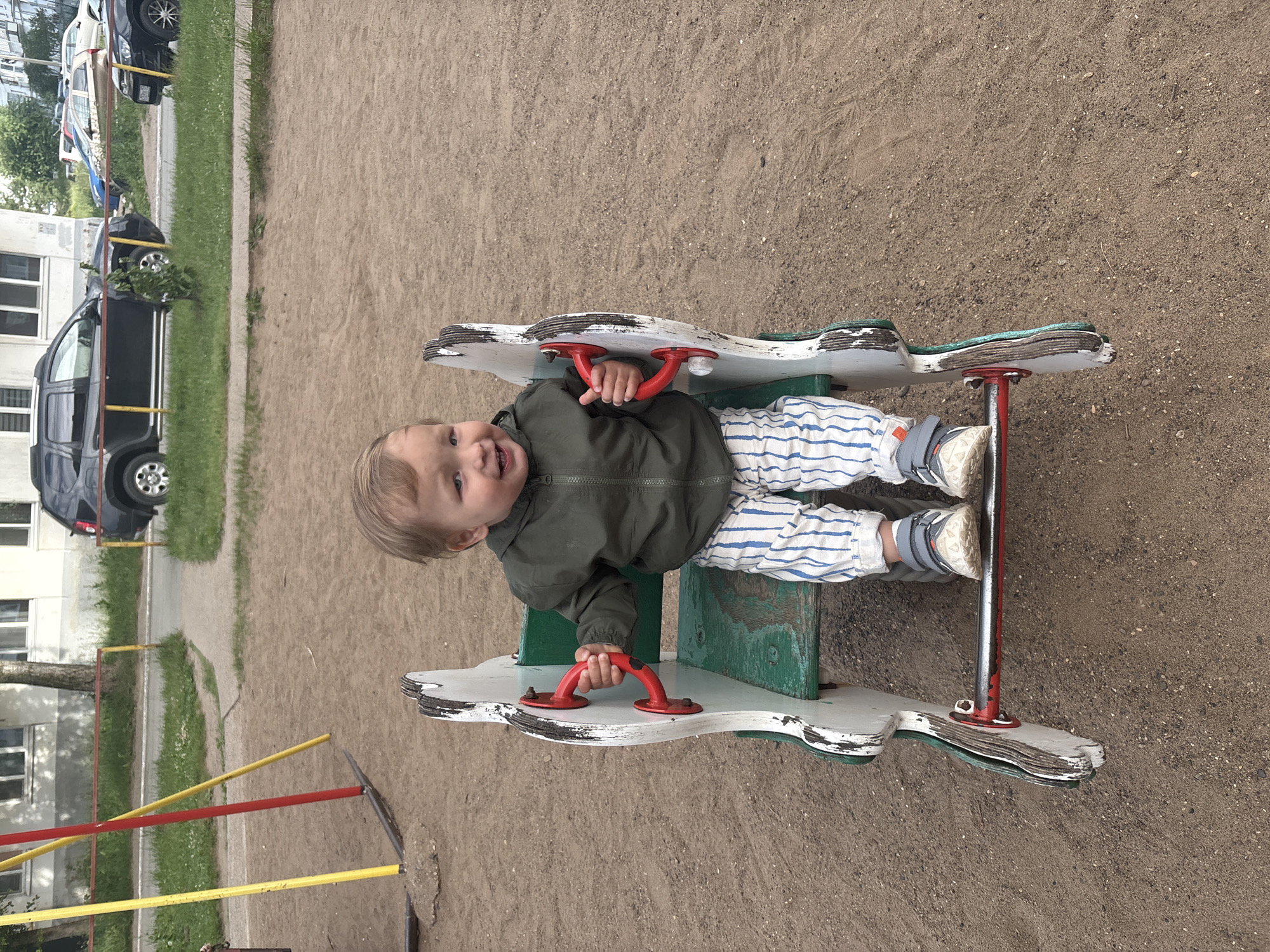 A young child, appearing to be between one and two years old, is seated on a worn, green, wooden seesaw-like playground toy. The child is wearing a dark green jacket, white pants with blue stripes, and grey sneakers with white soles. The child has blonde hair and is smiling, looking towards the camera and holding red handles on either side of the toy. The playground is covered in fine, brown sand. In the background, there are yellow and red poles of a swing set, a paved walkway, green grass, and a row of apartment buildings. Cars are visible parked on a street in front of the buildings, including a black SUV and a silver sedan. The scene appears to be a public playground in the city of Chaikovsky, Russia.