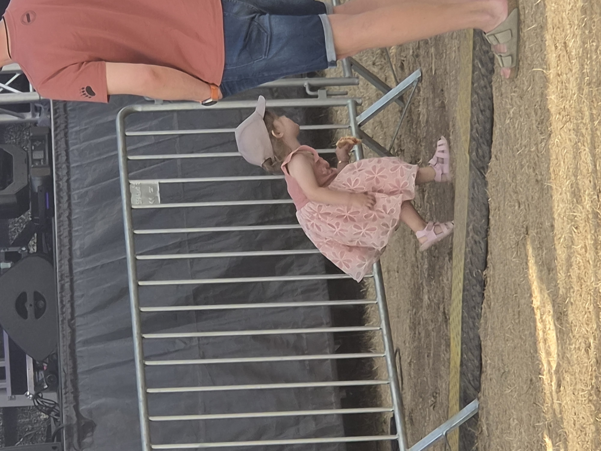 A young girl wearing a pink dress and a beige baseball cap is crouching near a metal barrier at an outdoor event in Sanem, Luxembourg. She is holding a piece of food in her right hand. To her right, an adult wearing a reddish-brown t-shirt and denim shorts stands partially in the frame. The background shows a stage with large speakers and a black tarp. The ground is covered in dry grass and dirt, with a yellow and black striped barrier visible on the ground.