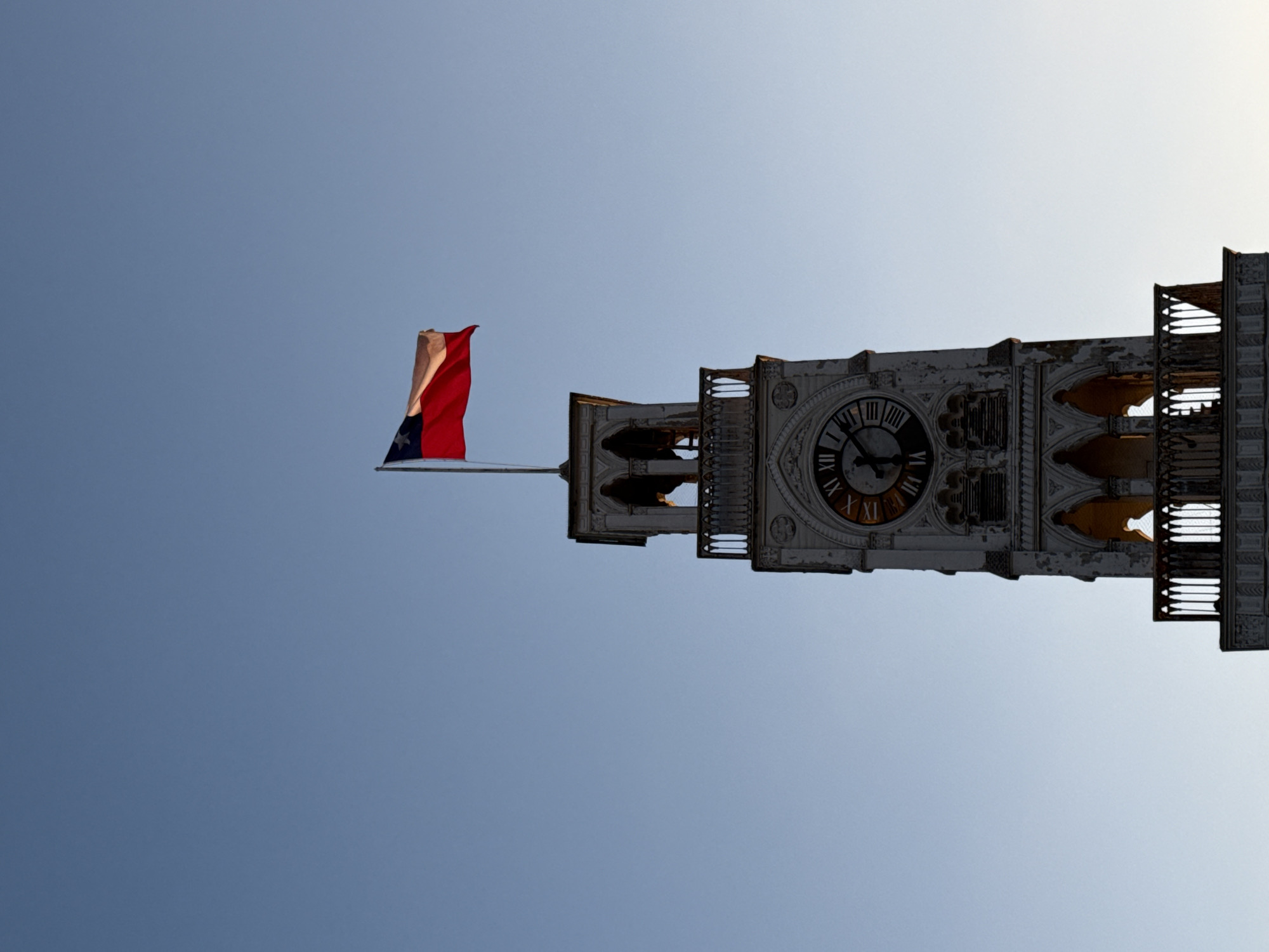 The image shows a close-up, low-angle shot of the clock tower in Iquique, Chile, from a distance, with a clear blue sky as the background. The tower is made of white stone and features intricate gothic architecture. At the very top of the tower, the Chilean flag is waving in the breeze. Below the flag, the tower has an open-air section with decorative railings and arched openings. A large clock face with Roman numerals is visible on the tower, indicating the time is approximately 10:50. The lower part of the tower features more arched architectural details and balconies. The overall impression is one of historical grandeur against a serene sky.