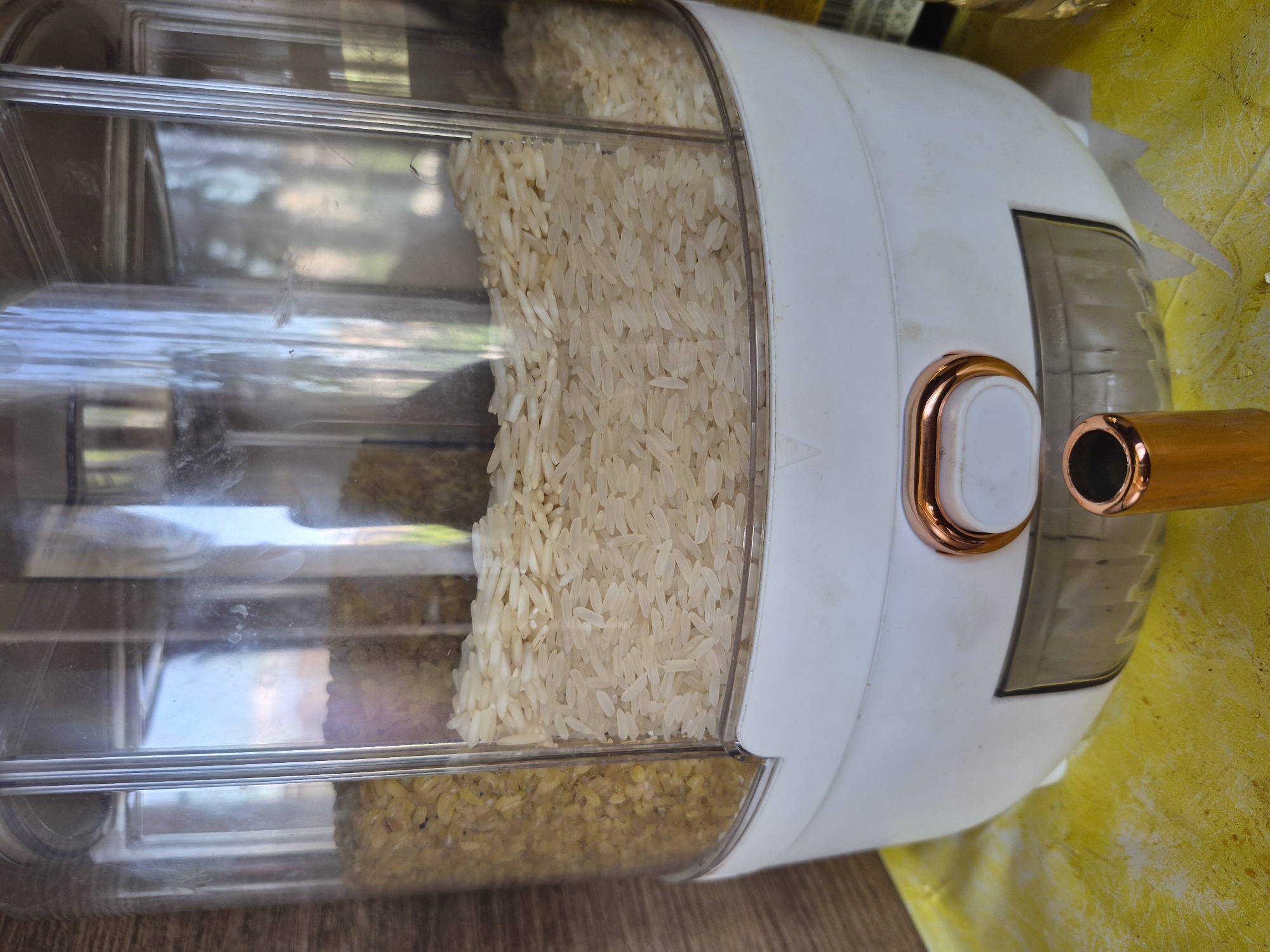 A modern, multi-compartment food dispenser, likely for grains, is depicted in a horizontal orientation. The dispenser features a predominantly white, opaque body contrasted with a large, transparent plastic front panel. Within this clear panel, the upper section is filled with a substantial quantity of light-colored, long-grain rice, appearing translucent. Below this, a separate compartment holds a different type of grain, which is smaller and yellowish, possibly bulgur or a different rice variety.
The dispenser is adorned with sleek, copper-colored accents. A central, rectangular mechanism, potentially a handle or a push-button for dispensing, is visible, along with a matching copper-colored spigot or handle protruding from the right side of the unit.
The dispenser rests on a light brown wooden surface. A bright yellow, possibly fabric, material is visible in the background to the right. Reflections on the clear plastic suggest an indoor setting with ambient light, indicating it's likely daytime. No specific activity beyond the storage of grains is occurring, and no people are present. There is no visible text on the dispenser. This image was captured in Yerevan, Armenia.