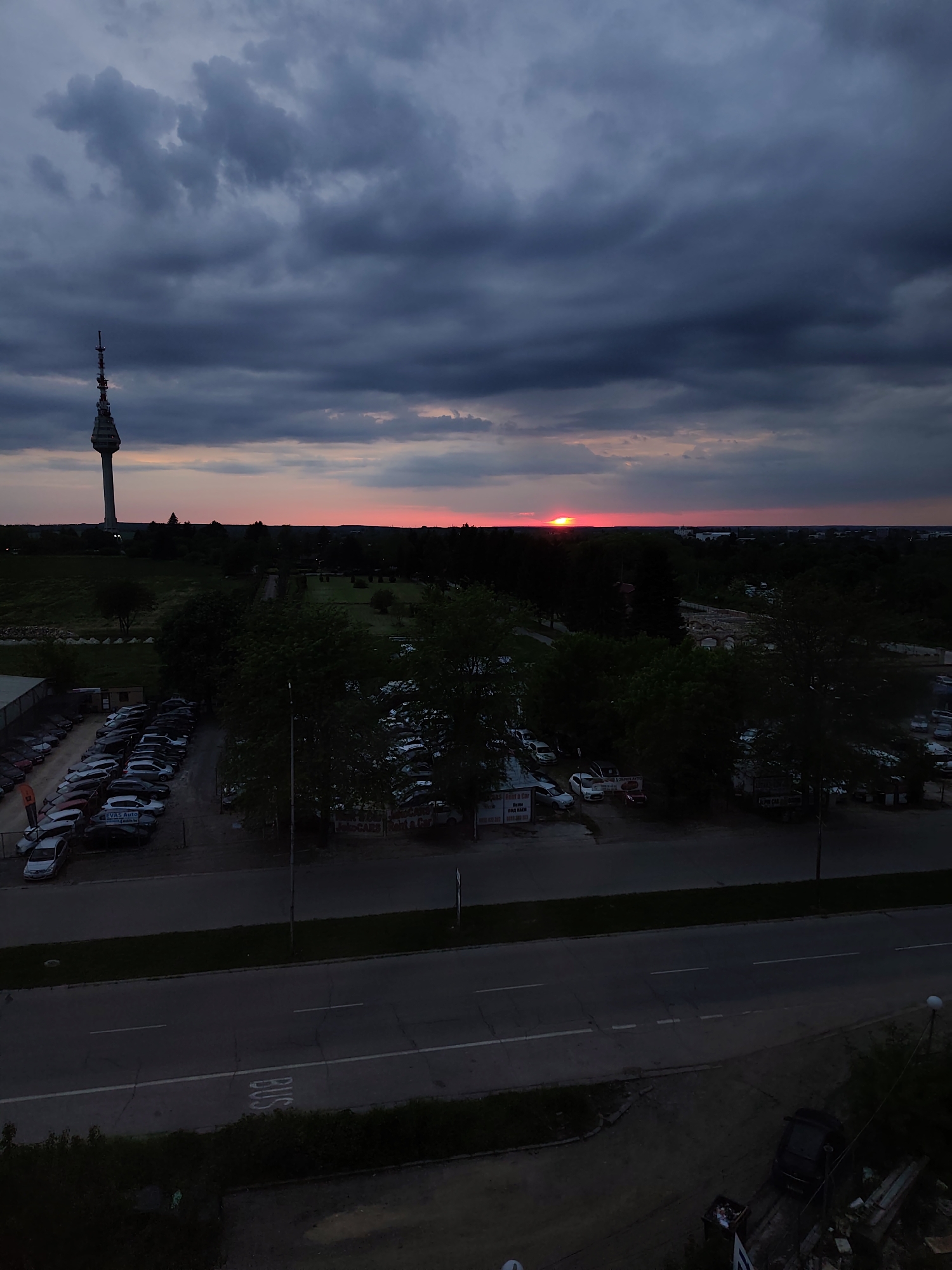 The image presents an elevated view of Dobrich, Bulgaria, at dusk or dawn. A tower stands prominently on the left horizon against a sky filled with dark, heavy clouds. A narrow band of red light, presumably from the setting or rising sun, breaks through the clouds on the horizon. Below, a line of trees separates a field from a lower level. There are several parking lots filled with cars on both sides of a street. The road is marked with lane lines and the word "BUS" painted on the surface. The foreground is dark, obscuring details.