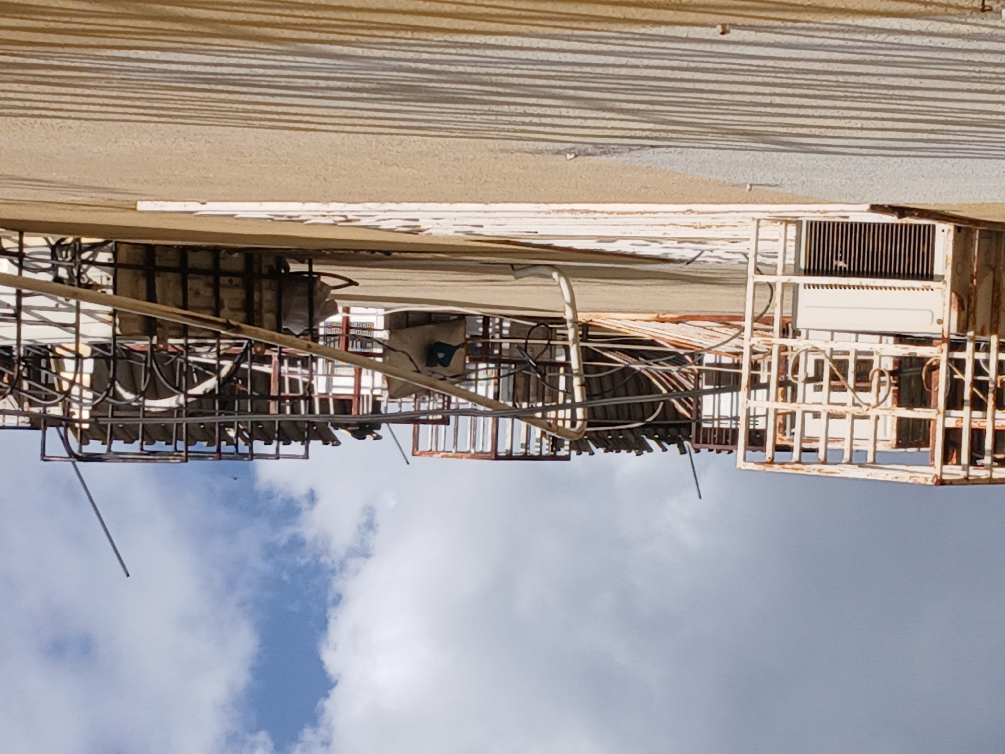 The image shows a building facade with multiple air conditioning units and metal framework against a backdrop of a cloudy sky. The location is Ciudad Guayana, Venezuela. The building appears to be weathered, with signs of rust on the metal structures and peeling paint on the building's surface. Various cables and conduits are visible, attached to the facade and framework. The sky is partly obscured by the building, revealing white clouds against a blue background.