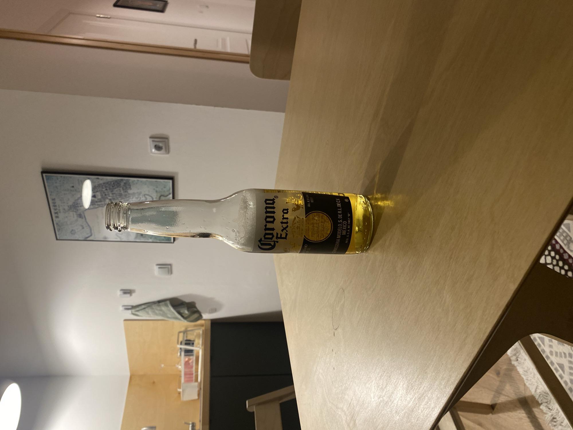 A nearly empty Corona Extra beer bottle lies horizontally on a tan-colored table. The bottle label is visible, containing brand and product information including "Corona Extra," "Alc. 4.5%," and "MEXICO." The background includes a wall with a framed picture, a light switch, and a piece of clothing or cloth hanging on the wall. A partial view of a kitchen or dining area is visible in the lower section of the image, including cabinets and possibly a small refrigerator. The scene appears to be indoors, possibly in an apartment or home. The image provides no information relating to the city of Izhevsk, Russia.
