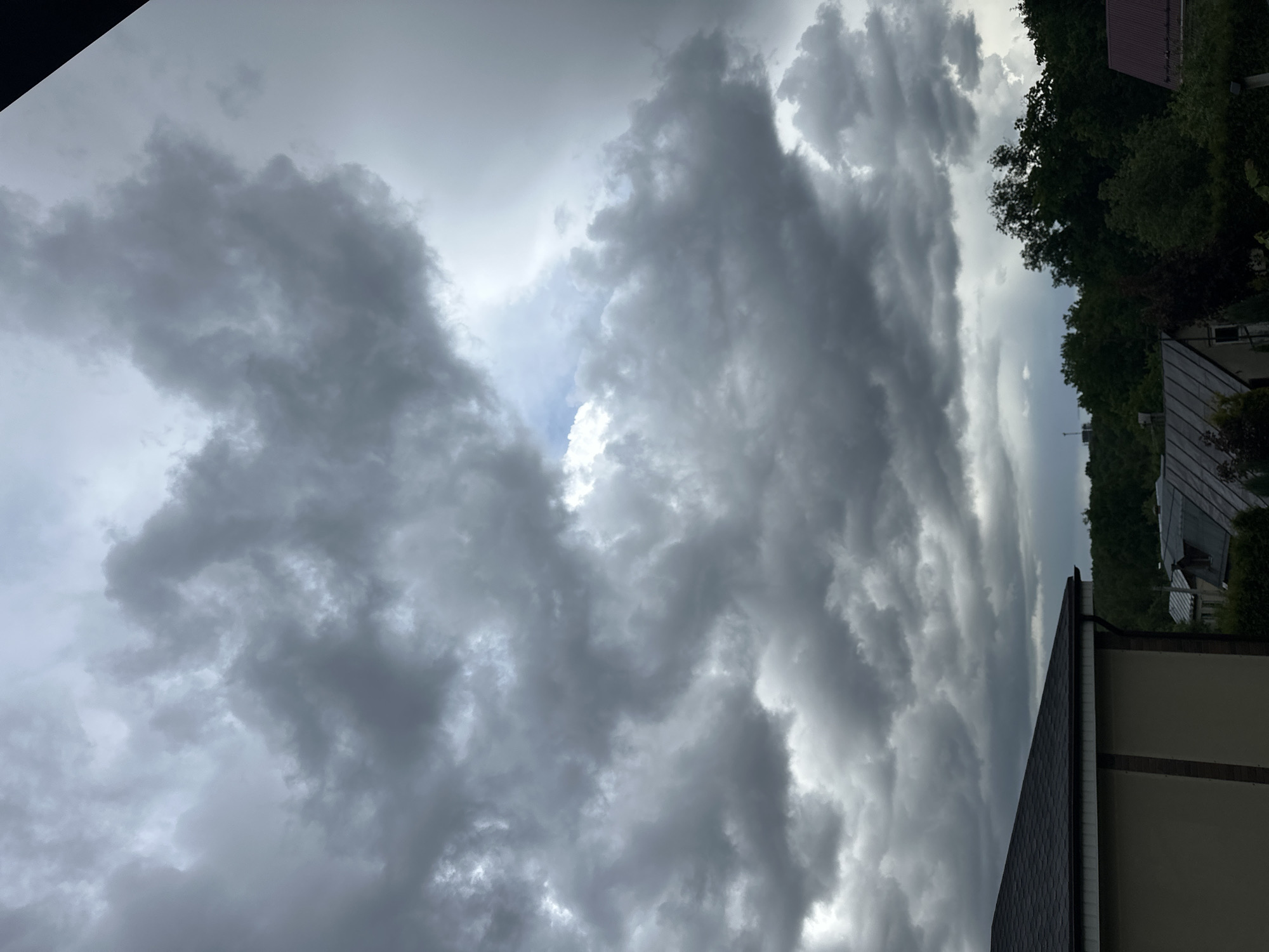 The image shows a view of a cloudy sky, possibly indicating an approaching storm. The clouds are a mix of gray and white, with some areas appearing denser and darker. A small patch of blue sky is visible through a gap in the clouds.
In the lower-right portion of the image, part of a building is visible, including a roof and wall. Some trees and other foliage can also be seen in the background.
Without more information, it is impossible to confirm the location of the image as being in Nizhny Novgorod, Russia. The image is a general representation of stormy weather.