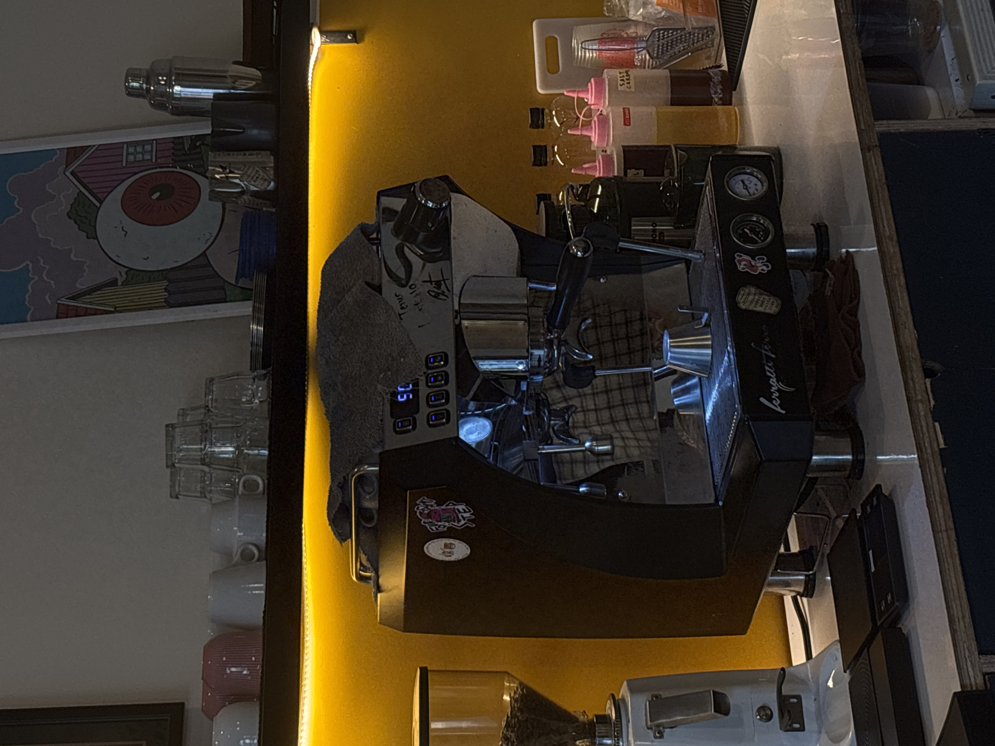 The image depicts a coffee machine situated on a countertop in what appears to be a cafe or coffee shop. The machine is dark-colored with chrome accents and has a digital display. To the left of the coffee machine is a coffee grinder, also predominantly white and chrome. Behind the coffee machine, a yellow wall is visible, adorned with a colorful framed artwork featuring a large eye. Several bottles of syrups and containers are lined up on the counter behind the coffee machine, along with a grater. In the background, a shelf holds various cups, both ceramic and glass. The lighting in the scene is dim, with a strip of yellow LED lights illuminating the area above the coffee machine and along the shelf. The overall impression is one of a functional, well-equipped coffee preparation area. This scene is located in the city of Semarang, Indonesia.