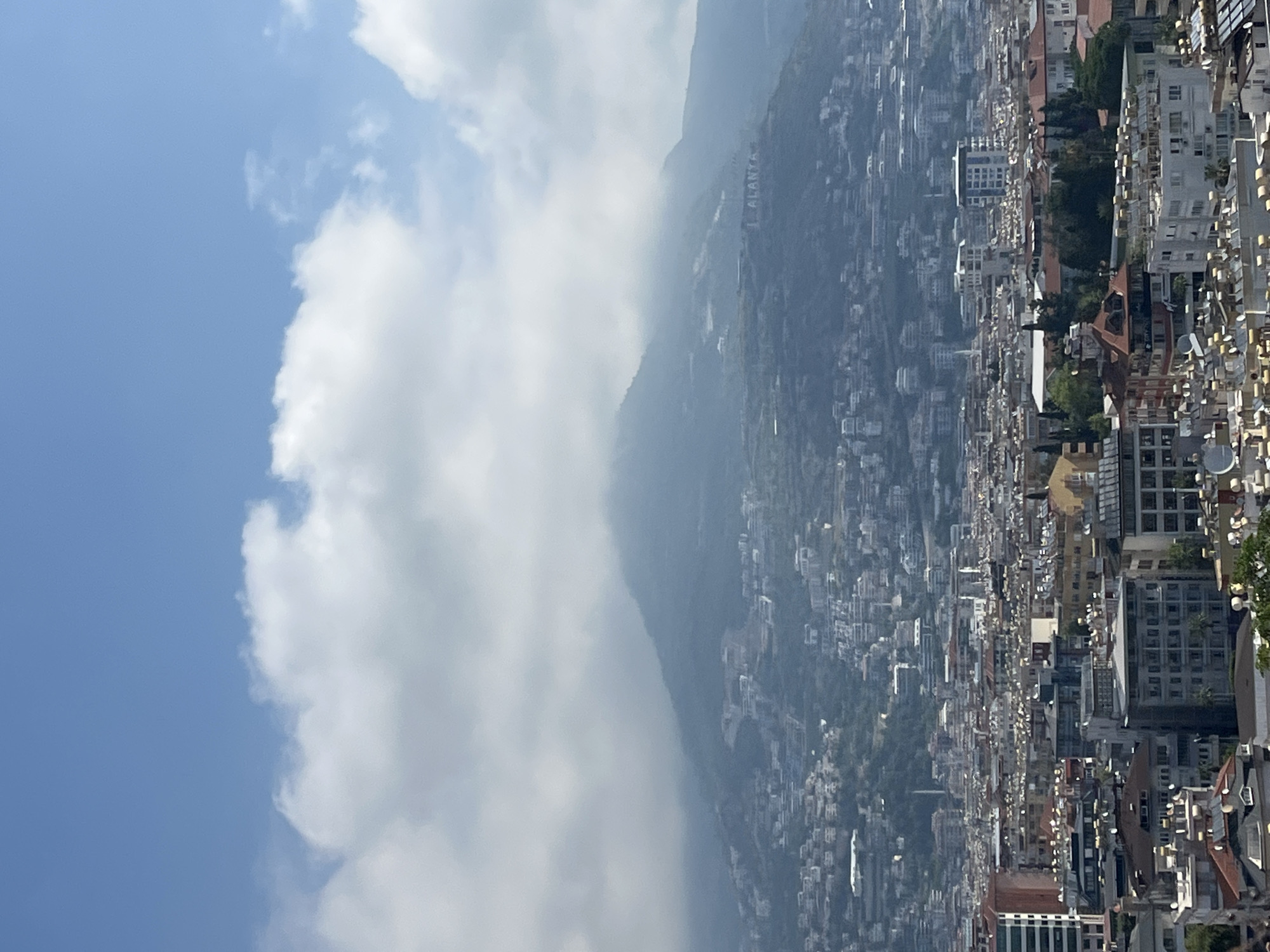 This media file presents an expansive, panoramic view of a densely populated city nestled at the base of a significant mountain range. The image is oriented vertically but depicts a horizontal landscape.
The foreground and mid-ground are dominated by the urban sprawl of the city, characterized by countless residential and commercial buildings. These structures vary in size and height, showing a mix of architectural styles, with many featuring light-colored facades and tiled roofs. Green patches of trees are visible amidst the buildings, particularly towards the mountain base.
Behind the city, a large, prominent mountain rises, its slopes covered in vegetation and partially obscured by a light atmospheric haze or mist. On this mountainside, clearly visible in large white letters, is the name "ALANYA". Further in the distance, additional mountain peaks can be discerned, fading into the background.
The upper portion of the image shows a clear blue sky, partially filled with large, white, cumulus clouds, especially on the left side, which also gently spill over the mountain. The lighting indicates it is daytime, likely under bright sunlight, contributing to the slight haze over the distant mountains. The overall scene suggests a vibrant coastal city integrated into a dramatic natural landscape.