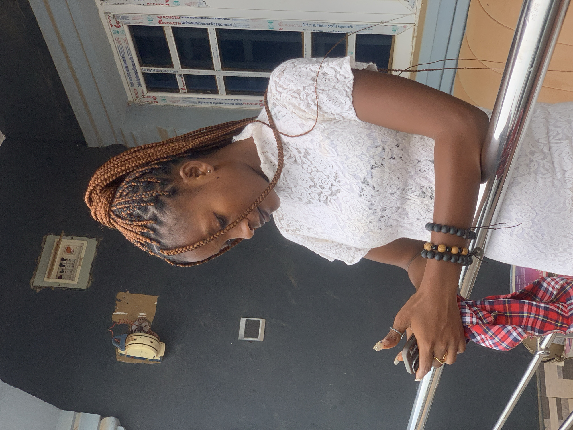 The image captures a young Black woman leaning against a silver railing outdoors, likely on a balcony or porch in Ikorodu, Nigeria. She has light brown braided hair, styled in cornrows, and wears a white lace dress. A small stud earring is visible in her left ear. Her right arm rests on the railing, showcasing multiple beaded bracelets (black and wooden) and rings on her fingers. She holds a phone in her right hand, partially covered by a red, white, and blue plaid fabric.
The woman is looking downwards, with a calm or contemplative expression. The background features a dark, possibly charcoal-grey, wall. A white-framed window with multiple panes is visible in the upper background. Below the window on the dark wall, there are two exposed electrical boxes or panels. The lighting suggests it is daytime.
Visible text on the window frame includes repeated branding such as "RONGTA," "Global aluminium profile exponential," and "Global aluminium picfile exponential." The railing also bears the text "SUS 304," indicating the type of stainless steel used.