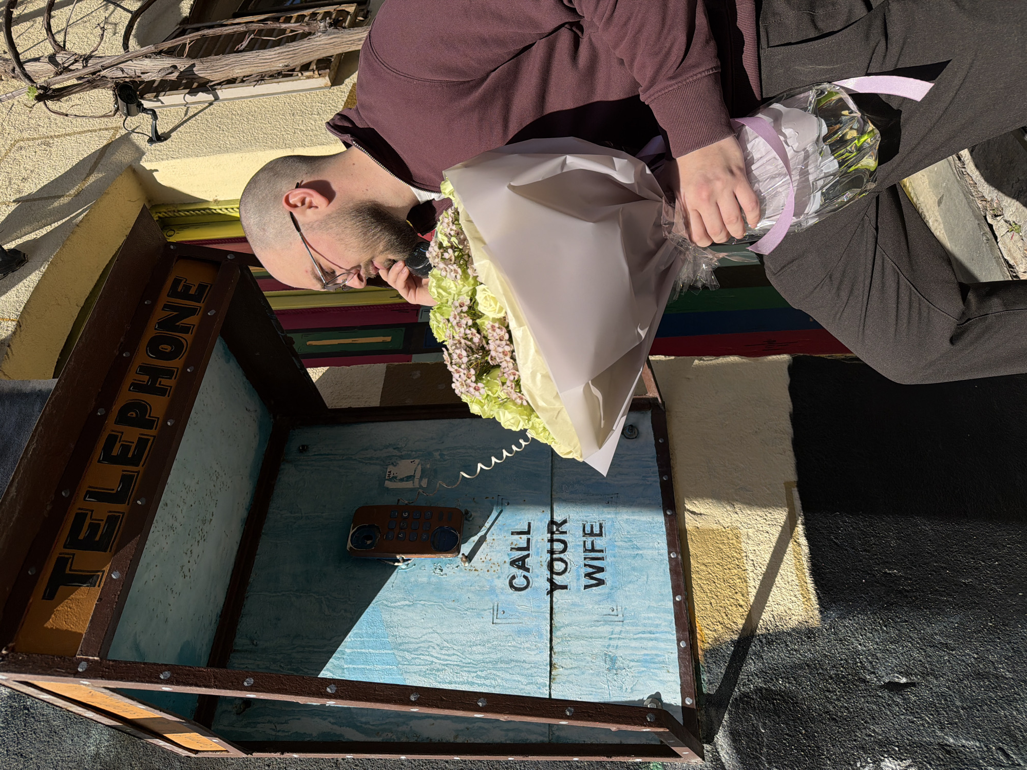 A man with a shaved head and glasses, wearing a maroon sweatshirt and black pants, is holding a bouquet of flowers wrapped in white and clear cellophane with a purple ribbon. He is pretending to talk on a telephone inside a wooden phone booth. The inside of the phone booth is painted light blue and has the words "CALL YOUR WIFE" painted in black. A small, brown, wooden phone is mounted inside. The exterior of the booth says "TELEPHONE" in yellow letters. The scene is outdoors, likely in Tbilisi, Georgia. The background includes a yellow building and a partly visible bicycle.