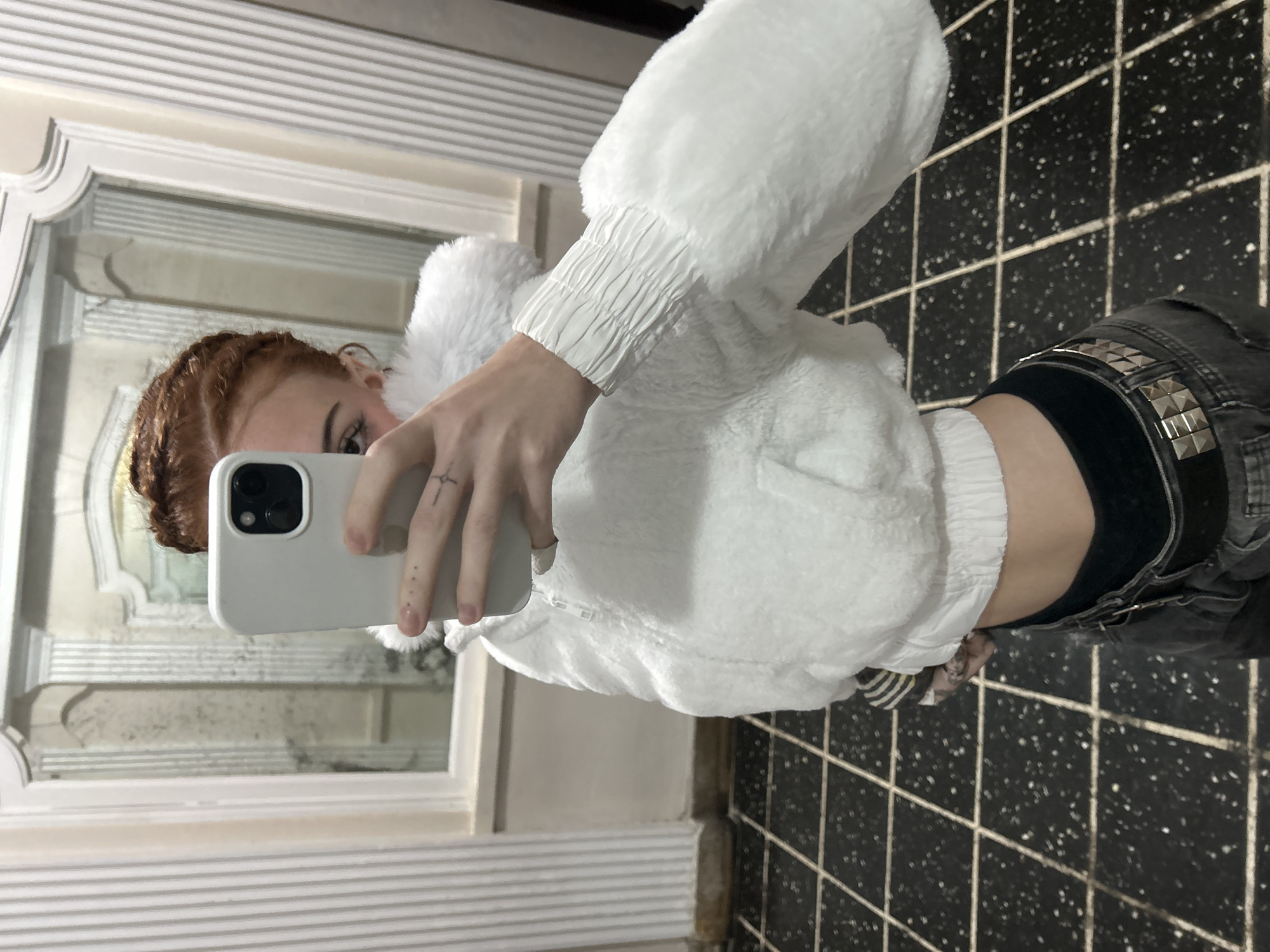 A reddish-haired person is taking a selfie in front of a mirror in what appears to be a bathroom. The mirror is encased in a white frame with horizontal lines. The person is wearing a white, furry jacket and black jeans with studded details on the waistband. The background consists of a tiled floor with a black and white grid pattern and a small portion of the bathroom wall. The location cannot be determined as Buenos Aires based on the image information provided.