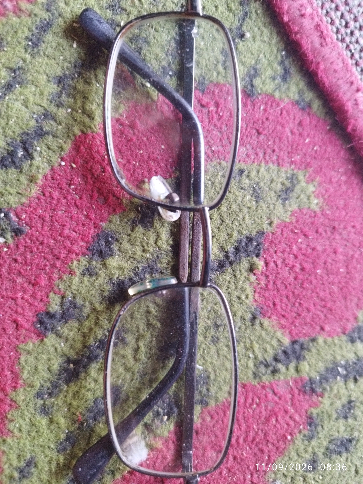 The image shows a pair of glasses lying on a patterned rug. The rug has red, green, and black stripes. The glasses are made of metal with clear lenses. One of the lenses is cracked. The timestamp on the image indicates it was taken on November 9, 2026, at 8:36 AM. The location is specified as Mojokerto, Indonesia.