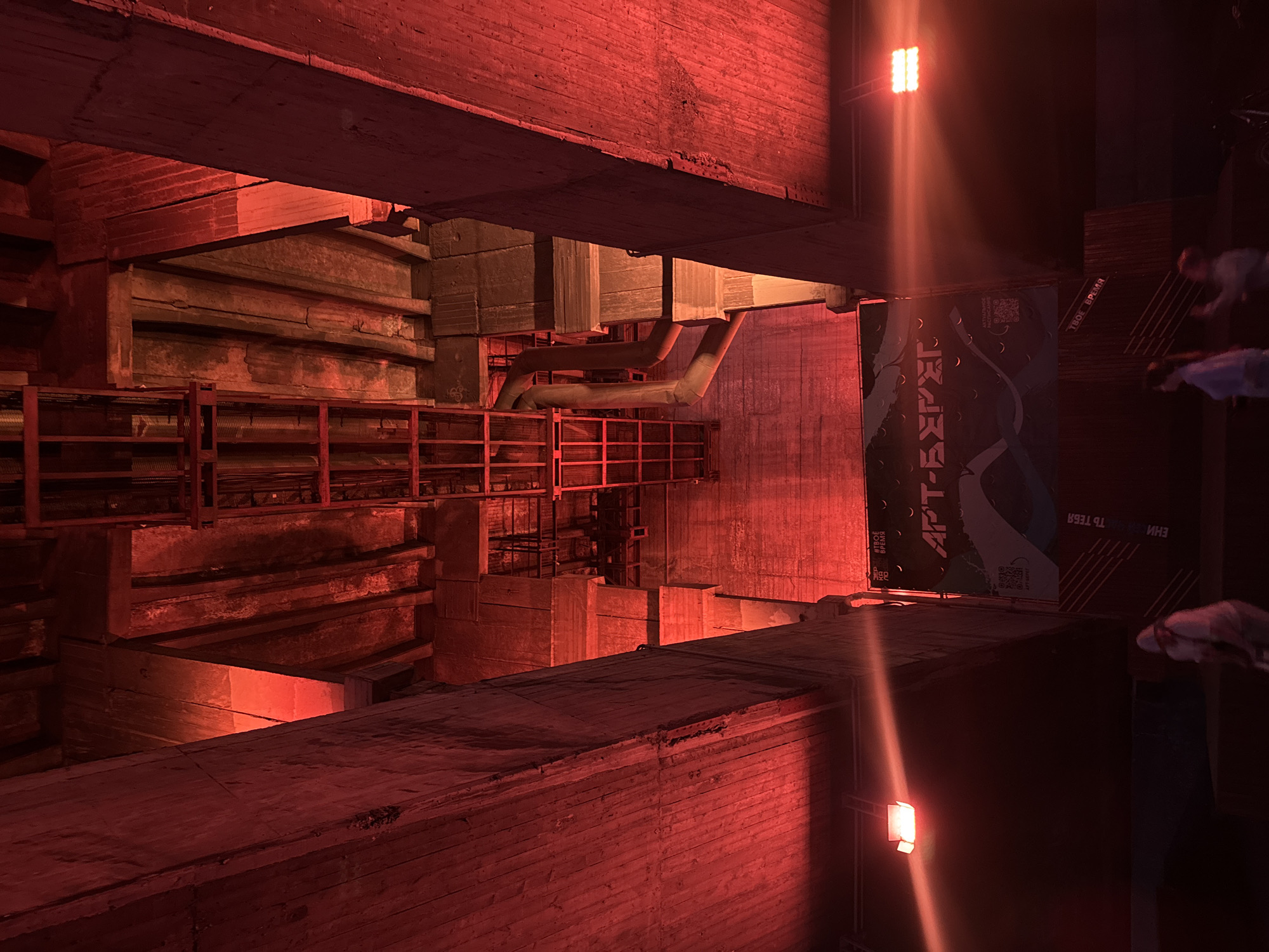 The image depicts the underside of a large concrete structure, possibly a bridge or overpass, in Krasnoyarsk, Russia. The scene is dramatically lit with red lights, casting deep shadows and highlighting the rough textures of the concrete and metalwork. In the background, there are visible pipes, scaffolding, and a large banner with Cyrillic text that reads "APT-БЕРЕГ" and possibly "ТВОЕ ВРЕМЯ". Three indistinct figures are visible in the foreground, walking or standing. One figure on the left is wearing a white hooded garment. Another figure in the center is wearing a light-colored top. A third figure on the right appears to be partially visible. The overall impression is one of an industrial or artistic space, possibly an event venue or a public art installation, illuminated for nighttime viewing.