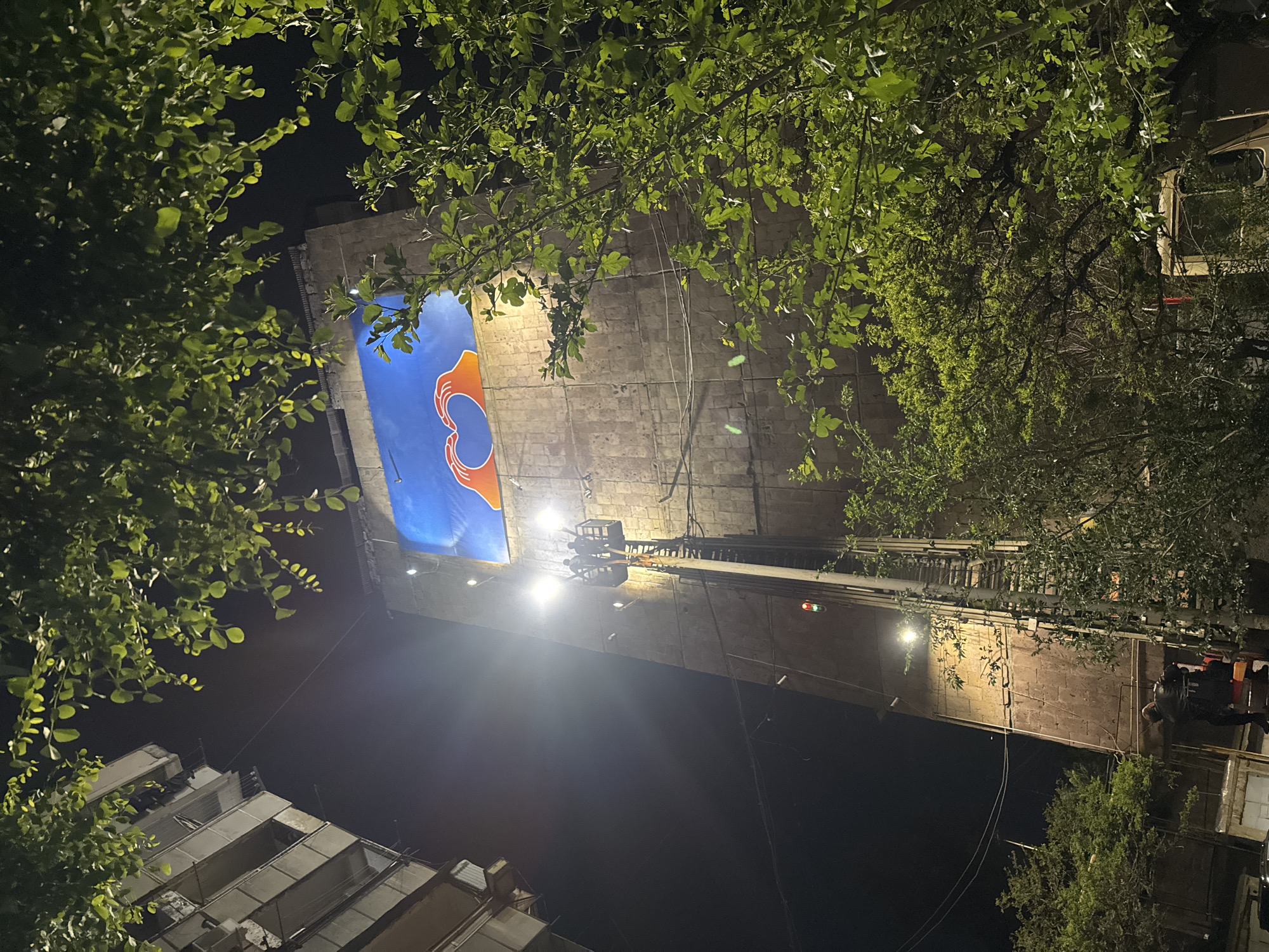 The image shows a night scene in Yerevan, Armenia, focused on a large building with a mural. The mural features a blue background with an orange design of two hands forming a heart around a circle. A crane extends towards the mural, possibly for maintenance or installation. Lush foliage from trees frames the top and sides of the building, partially obscuring the view. In the lower left, a portion of an adjacent building is visible. The lighting is bright, particularly around the crane and the building with the mural, contrasting with the darkness of the night sky.