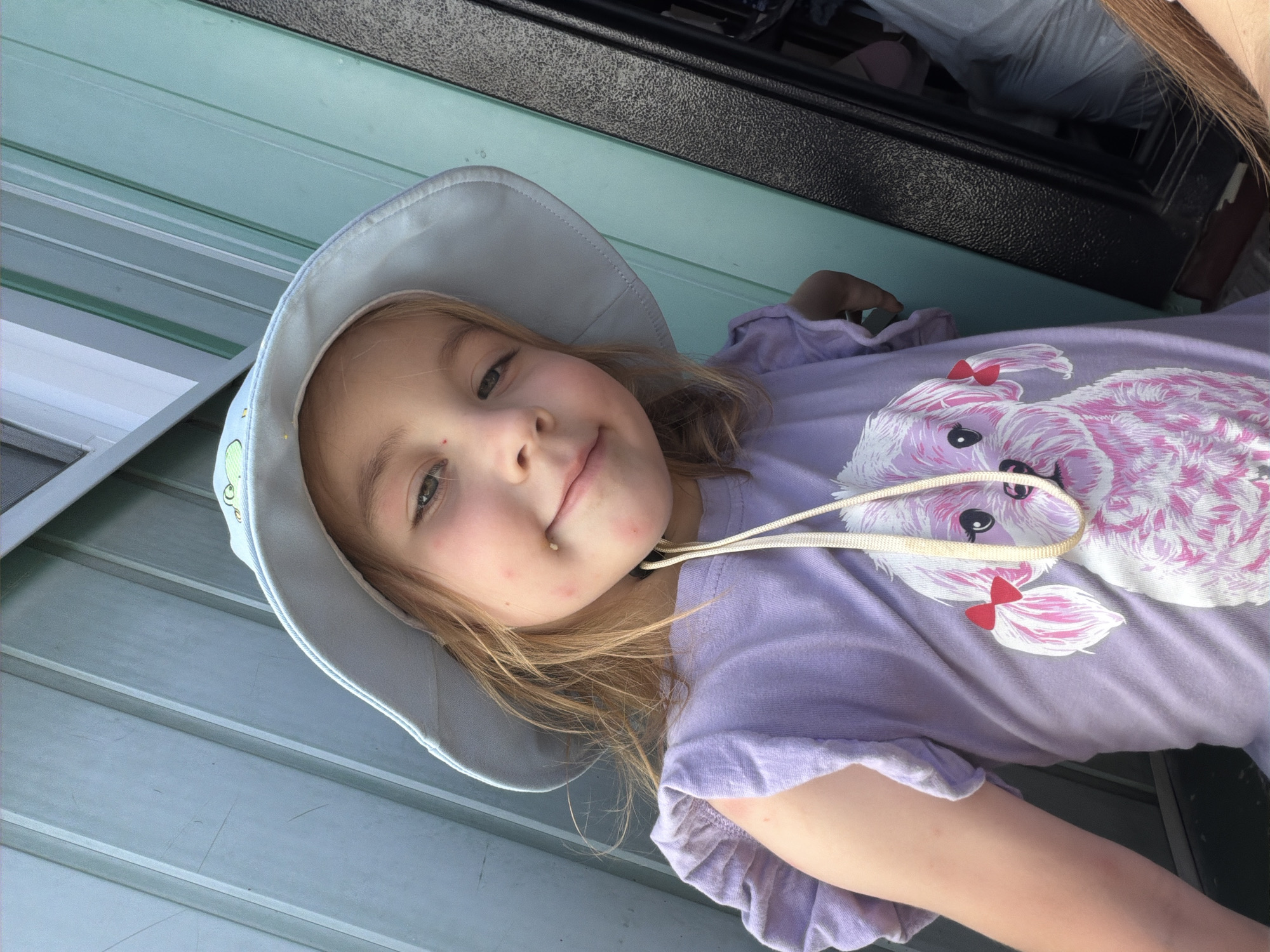 A young girl, wearing a light blue sun hat and a lavender t-shirt with a pink and white dog graphic, is captured in a close-up, selfie-style shot. She has blonde hair and appears to have some red marks on her face. The background consists of a light blue corrugated metal wall and a dark, textured surface, possibly a door or a cabinet. The image is taken outdoors, suggesting a residential or recreational area. There is no specific indication of the city of Tambov, Russia, in the visual information provided.