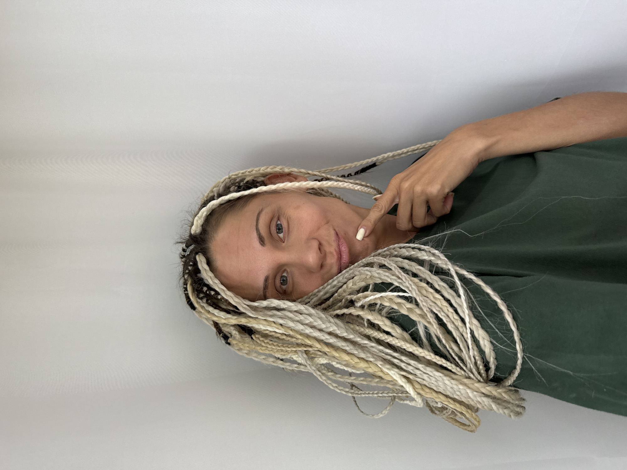 A woman with blonde and brown dreadlocks stands against a white backdrop, looking at the camera with a slight smile. She is wearing a dark green t-shirt and has her right index finger touching her chin. The image does not contain enough information to determine the location within Rusanovo, Russia.