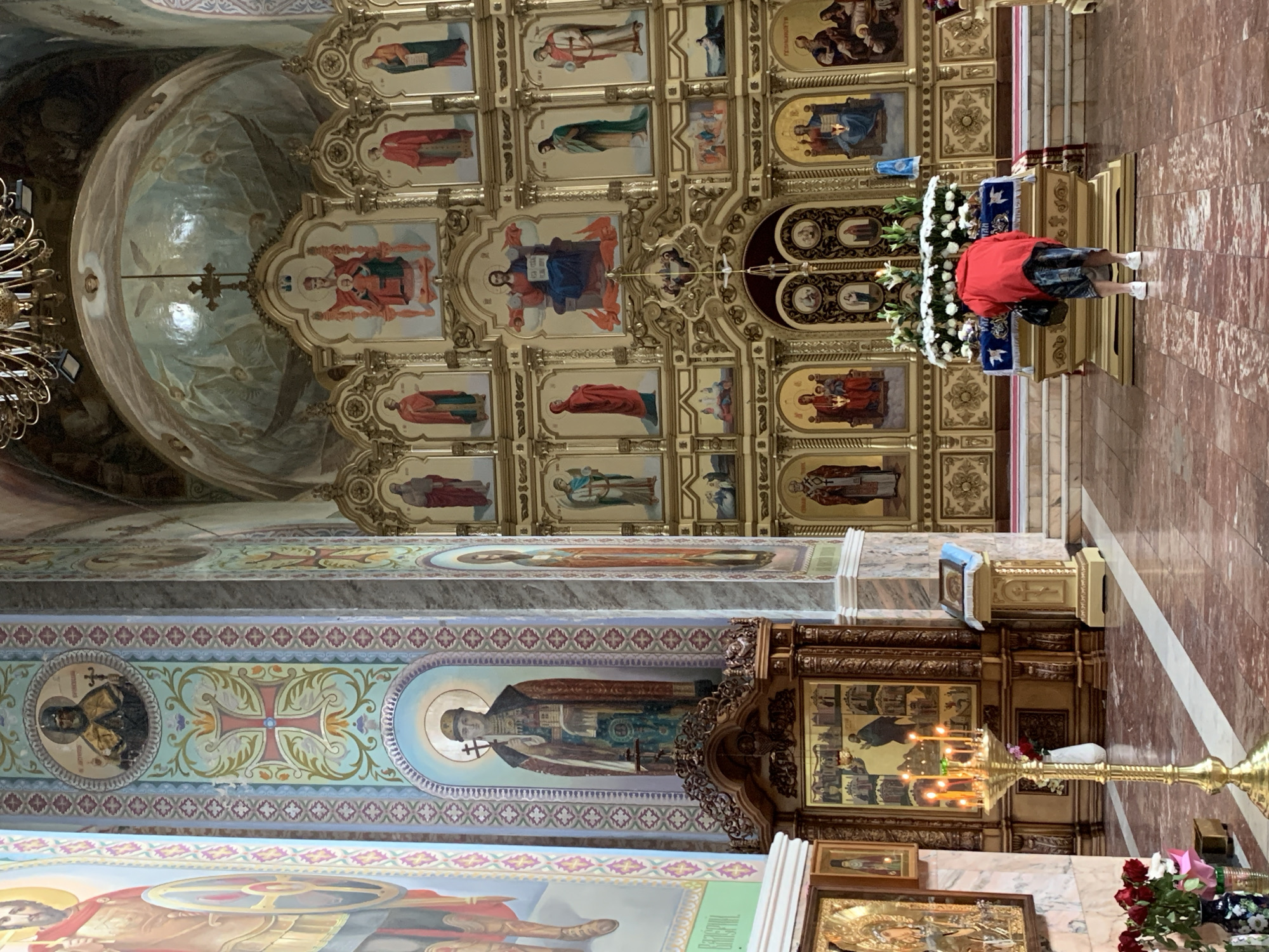 The image depicts the interior of a Russian Orthodox church, specifically the Cathedral of the Nativity of the Blessed Virgin Mary in Prokopyevsk, Russia. The scene is dominated by an ornate golden iconostasis, a wall of icons separating the nave from the sanctuary, adorned with numerous religious paintings. In the foreground, a woman wearing a red jacket and a patterned skirt is seen from behind, bent over a relic or altar decorated with white flowers. She is facing the iconostasis, with her hands near the floral arrangement. To the left, a tall brass candelabrum with lit candles stands on the marble floor. The walls and ceiling are also decorated with religious frescoes. The overall atmosphere is one of solemnity and devotion.