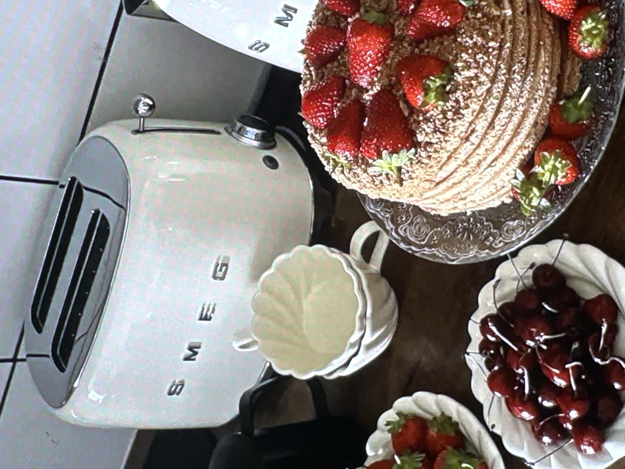 The image shows a still life arrangement featuring a Smeg toaster, a white scalloped cup, a cake decorated with strawberries, and bowls of cherries and strawberries. The Smeg toaster is positioned on the left, with the brand name clearly visible. The cake, adorned with fresh strawberries, sits on a glass cake stand. A white scalloped cup is placed near the toaster. Bowls containing cherries and strawberries are arranged around the cake. The image does not provide enough information to identify the specific location in Yerevan, Armenia.