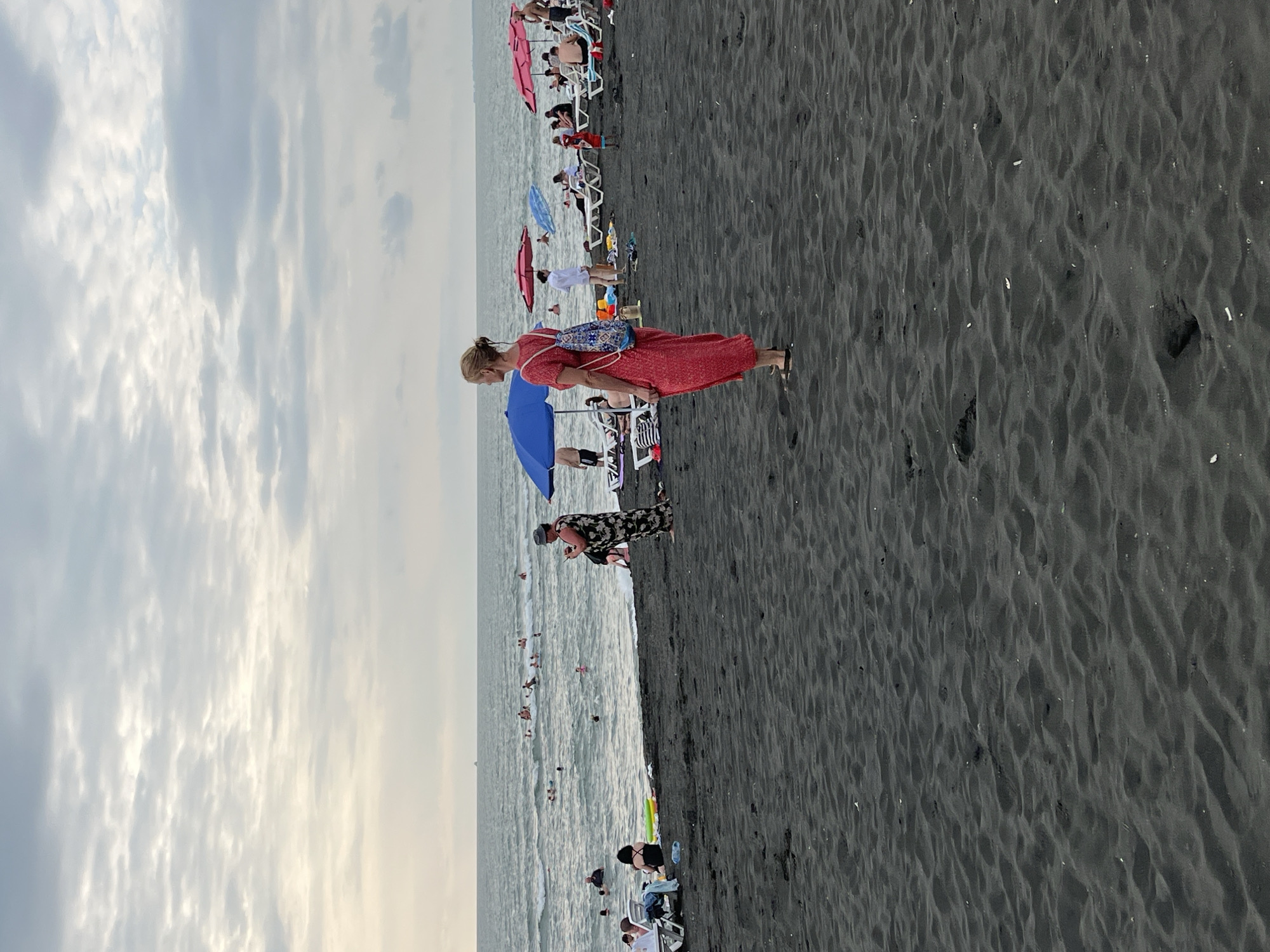 The image depicts a crowded beach scene in Ureki, Georgia, characterized by its distinctive black sand. The sky is overcast with clouds. In the foreground, the black sand is rippled and textured. A woman in a long, red dress and a backpack stands facing away from the camera, looking towards the sea.
Further back on the beach, several people are visible. Some are seated on lounge chairs, while others are standing or walking. Beach umbrellas, predominantly blue and pink, dot the scene, providing shade. A woman in a patterned dress is standing near the water's edge, holding a phone.
In the sea, numerous people are engaged in various water activities, including swimming and playing in the waves. The water is a muted grey-blue, reflecting the cloudy sky. The overall impression is a lively, somewhat overcast day at a popular seaside resort.