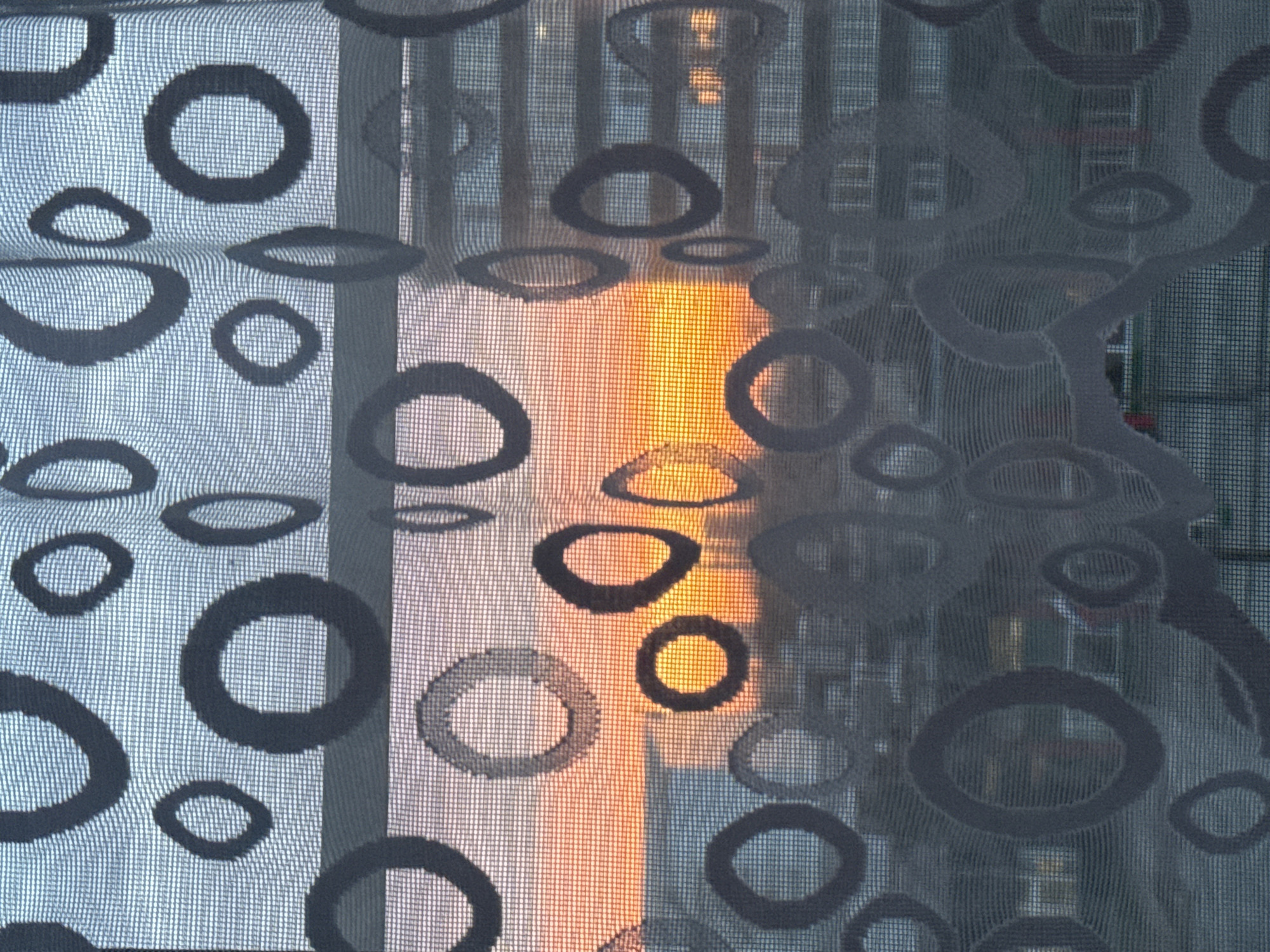 The image shows a window curtain with a circular pattern, partially obscuring a view of buildings, likely located in an urban setting. The lighting suggests it might be either sunset or sunrise, with a warm glow filtering through the fabric. The patterned curtain features a repeat of irregularly shaped circles in black or gray tones. The background appears to be of dense urban development; therefore it is unlikely the location is the city of Saint Petersburg, Russia.