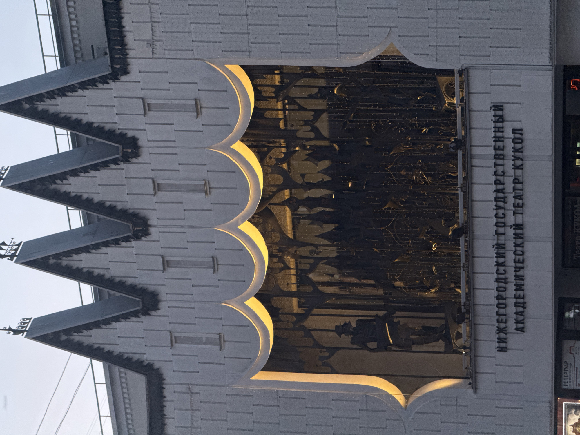 The image shows a building facade, identified by text in Russian as the Nizhny Novgorod State Academic Puppet Theatre. The architecture features a decorative pointed roofline and scalloped trim around a central window. The window displays a dark, detailed scene or sculpture with figures, possibly representing characters from puppet shows. The building is constructed from light-colored bricks or blocks. A poster or display case with additional information is visible on the right side of the image.
