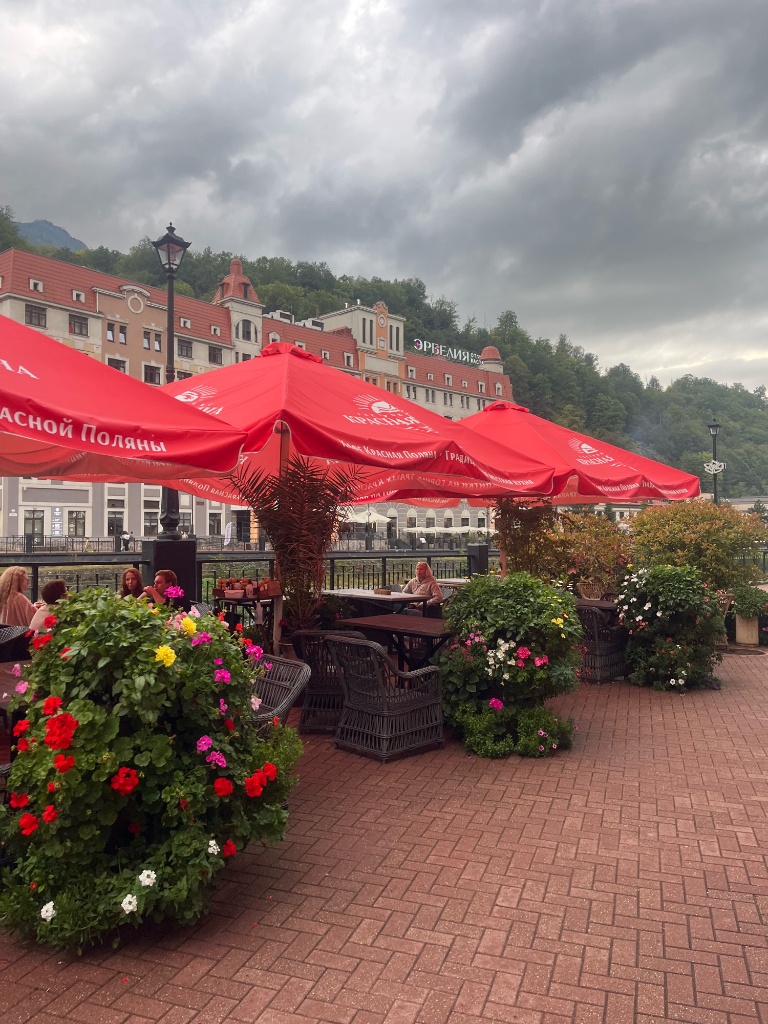 The image depicts an outdoor seating area of a cafe or restaurant in Estosadok, Russia, situated along a waterfront. The scene is dominated by large, vibrant red umbrellas providing shade over tables and chairs. Several patrons are seated at the tables, some engaged in conversation, others appearing to be dining.
In the foreground, abundant floral arrangements in large planters, filled with red, pink, yellow, and white flowers, add a splash of color to the brick-paved patio. Wicker chairs and tables are strategically placed throughout the seating area.
The background reveals a multi-story building with a distinctive architectural style, featuring red-tiled roofs and beige facades, consistent with buildings in resort towns. Behind the building, a lush green hillside covered with trees slopes upwards, under a dramatic, cloudy sky. The overall atmosphere suggests a relaxed, outdoor dining experience in a scenic mountain resort setting. The sign on the building above the umbrellas reads "ЭРБЕЛИЯ", which translates to "Erbelia".
