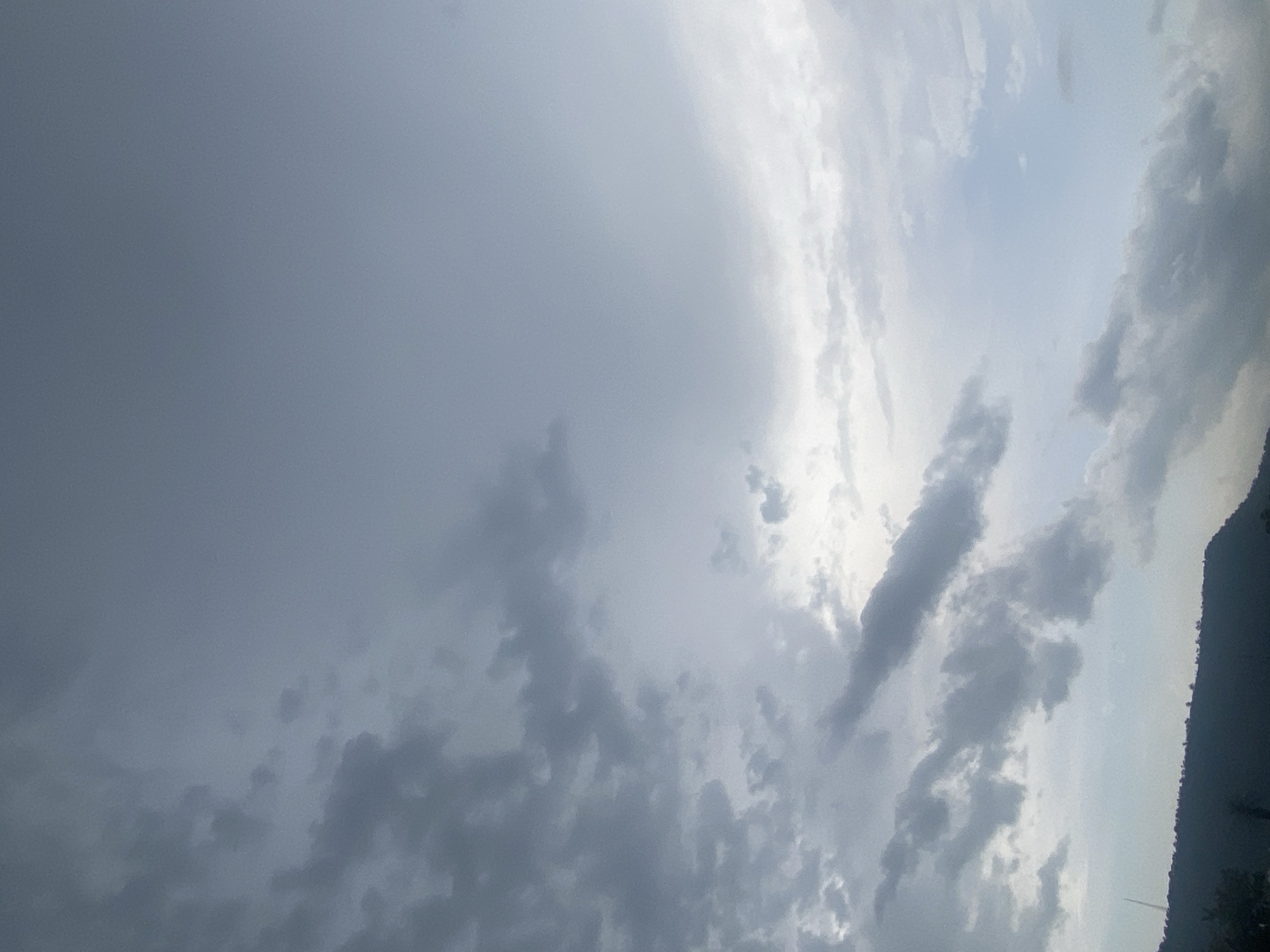 The image displays a wide expanse of sky dominated by diverse cloud formations. The color palette primarily consists of various shades of grey and white, suggesting an overcast or partly cloudy condition. On the left side of the frame, denser, dark grey clouds are prominent, indicating significant cloud cover. As the gaze moves towards the right, the clouds appear lighter and more fragmented, revealing brighter sections of the sky, likely where sunlight is either breaking through or diffused. The illumination source seems to originate from the upper right, casting bright white highlights on the edges of some clouds.
Along the far-right edge of the image, a dark, undulating silhouette resembling a mountain range or a series of hills is visible, rising against the brighter sky. This element firmly places the scene in a natural, outdoor landscape. There are no identifiable people, animals, or man-made objects distinctly visible within the frame.
The overall atmosphere conveys natural weather patterns, possibly hinting at an approaching or receding rain event given the blend of dark and bright cloud masses. The subdued lighting indicates either an early morning, late afternoon, or a heavily clouded daytime. No text is visible in the media. The landscape with hills is consistent with the geographical context of Lokoja, Nigeria.