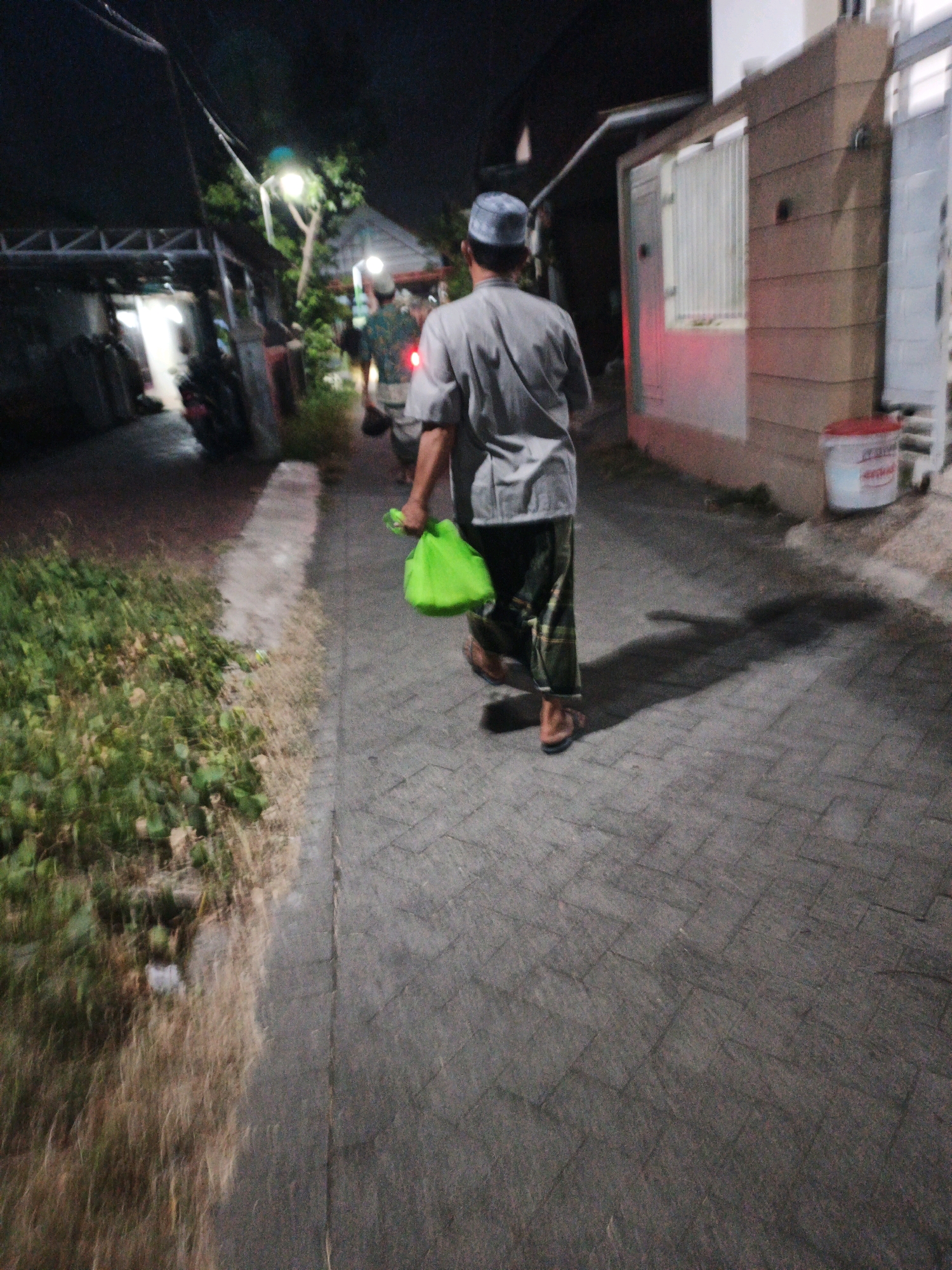 In Ketegan, Indonesia, at night, a person walks away from the camera down a paved pathway. The person is wearing a light gray shirt, dark patterned pants, a light blue cap, and brown sandals. They are carrying a bright green bag in their right hand. The pathway is bordered on the left by overgrown vegetation and on the right by a building with a light-colored exterior and a window with vertical blinds. A red and white cylindrical object, possibly a bucket, is visible near the building. The scene is illuminated by artificial lights, casting shadows on the ground. In the background, another person in a green patterned shirt is walking ahead, and there are structures suggesting residential or commercial buildings.