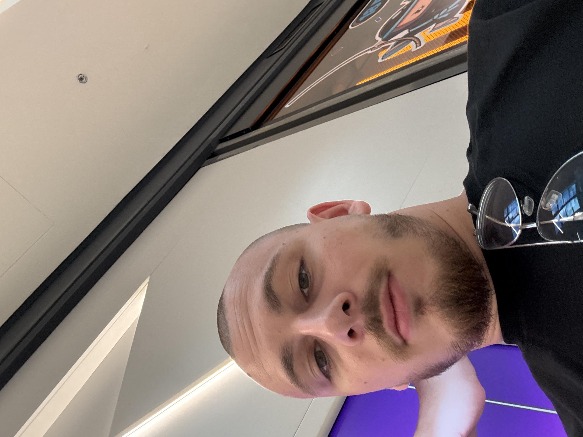 A selfie taken indoors. The subject is a man with a closely shaved head and a beard. He is wearing a black shirt and eyeglasses are hooked onto the collar. He is standing beneath a ceiling with recessed lighting and a possible decorative advertisement or sign above him. There is a purple surface or screen visible below him. A precise location within Burgas, Bulgaria cannot be determined from the image.