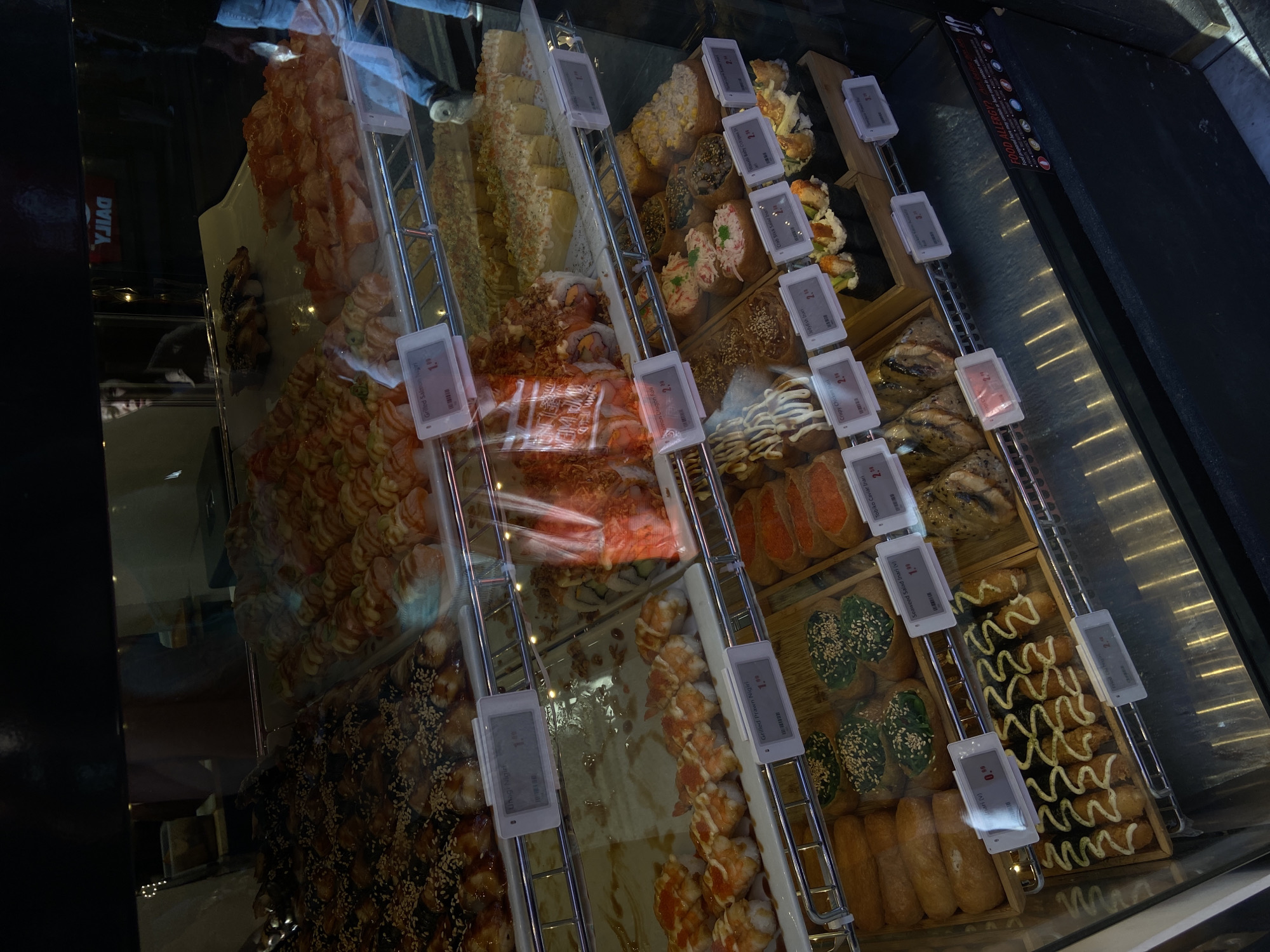 This is an indoor close-up shot of a display case filled with various types of sushi, likely located in a food establishment. The sushi is arranged in rows, with each type having its own section. There are small price tags with descriptions in English in front of each section.
Some of the types of sushi visible include:
* Grilled Salmon Nigiri
* Salmon Dragon Roll
* Tobiko Caviar Inari
* Crispy Chicken Inari
Other descriptions visible are "Unagi Nigiri," "Inari (v)," "Seaweed Salad Inari (v)", "Grilled Prawn Nigiri", "Jellyfish Inari", and "Crab Stick Salad Inan."
The display case is likely located in a store in London, United Kingdom, given the presence of "YJIAG" on the case's side, which is "GAIJY" spelled backward, and the high popularity of sushi in London. The overall lighting is bright and intended to showcase the food.