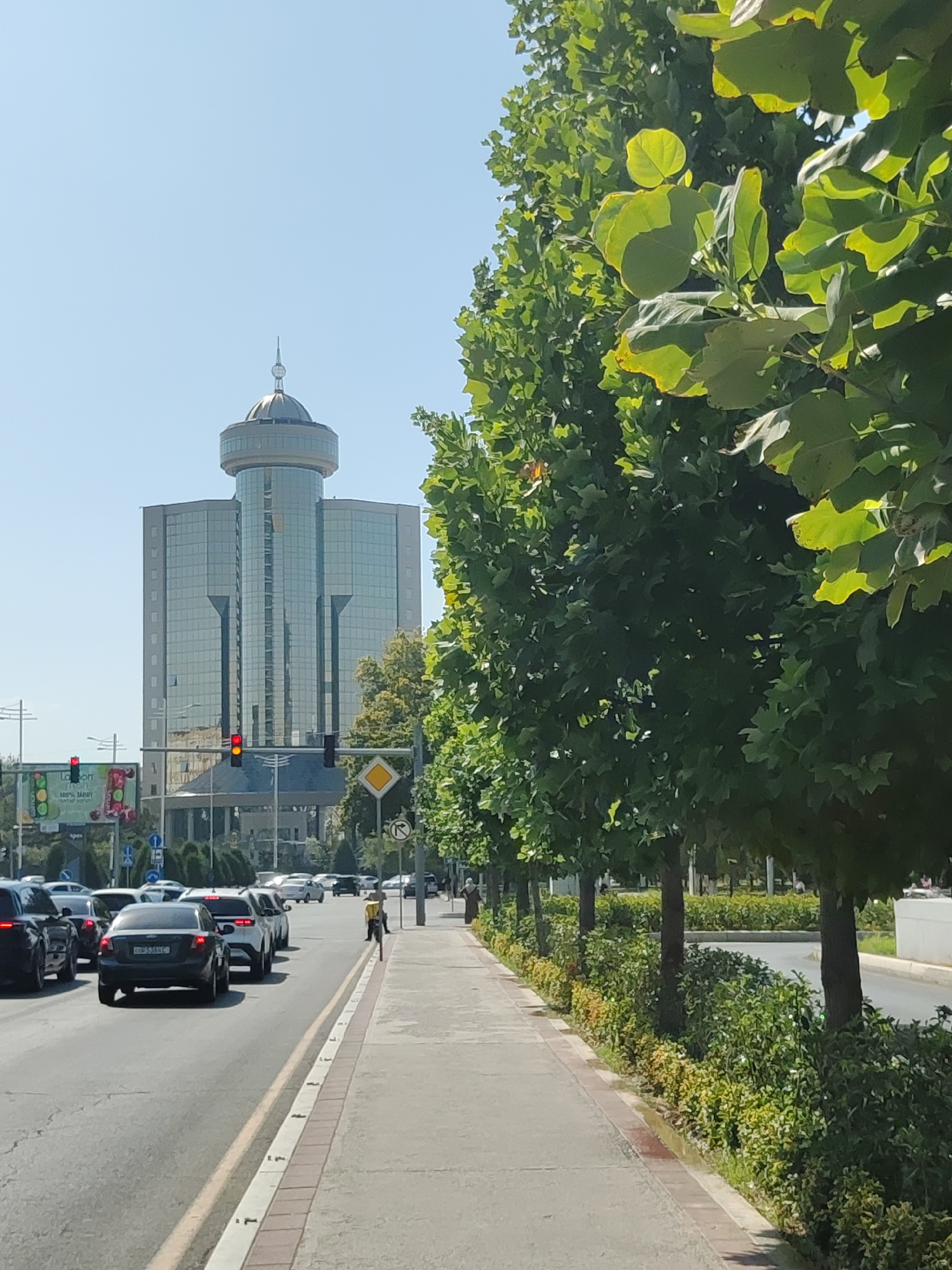 A modern glass skyscraper with a domed top stands on the left side of the image, reflecting the clear blue sky. The building is situated in Tashkent, Uzbekistan, likely a business district given its architecture. A tree-lined sidewalk runs along the right side of a busy street, with traffic moving in the foreground. Cars are stopped at a red traffic light. Several people are visible, including a pedestrian walking on the sidewalk and drivers inside their vehicles. The trees are lush green, suggesting a warm season. The scene captures a typical urban environment with a blend of modern architecture and natural elements.