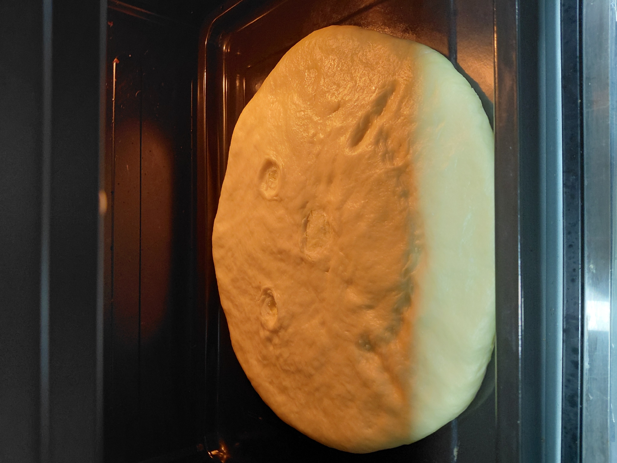 A raw, round loaf of dough is positioned on a baking tray inside an oven. The dough has indentations that resemble a face, with two circular depressions for eyes and a larger, curved depression for a mouth. The top portion of the dough is illuminated by the oven light, giving it a golden-brown hue, while the bottom portion remains a lighter, uncooked yellow. The baking tray is dark-colored and rests on oven racks. The background of the oven interior is dark and slightly reflective. The image does not contain any information about the city of Encarnación, Paraguay.