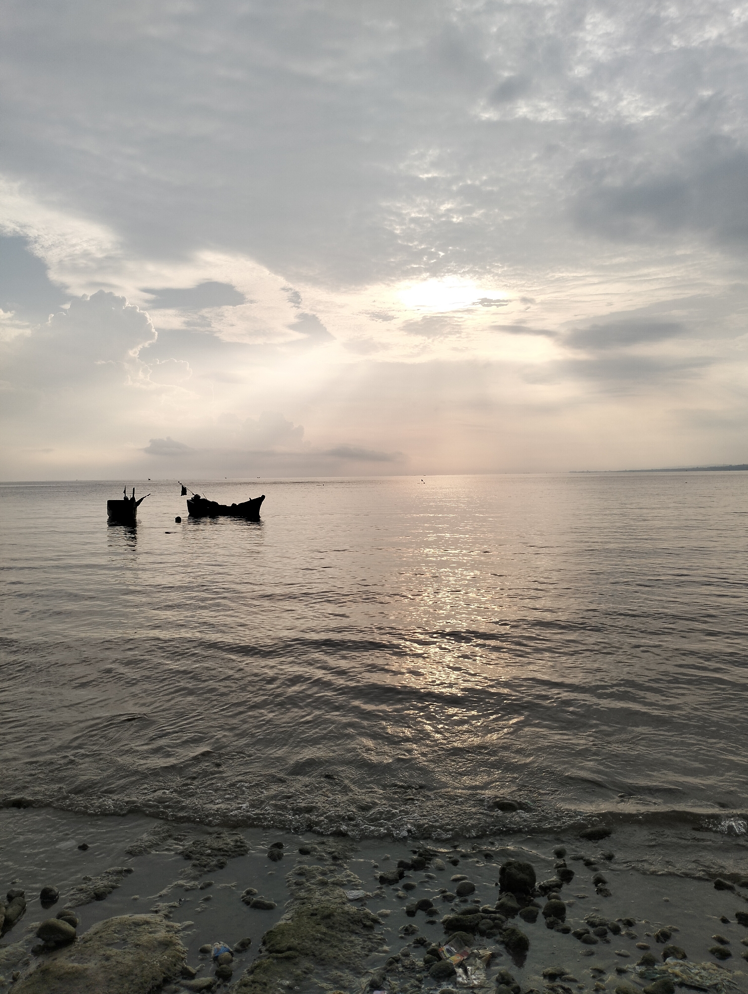 The image shows a seascape in Lhokmanee, Indonesia. Two dark silhouettes of boats are visible in the water. The water is calm with gentle waves, reflecting sunlight. The foreground shows a beach area with rocks and debris. The sky is overcast with patches of sunlight breaking through the clouds. No people are visible.