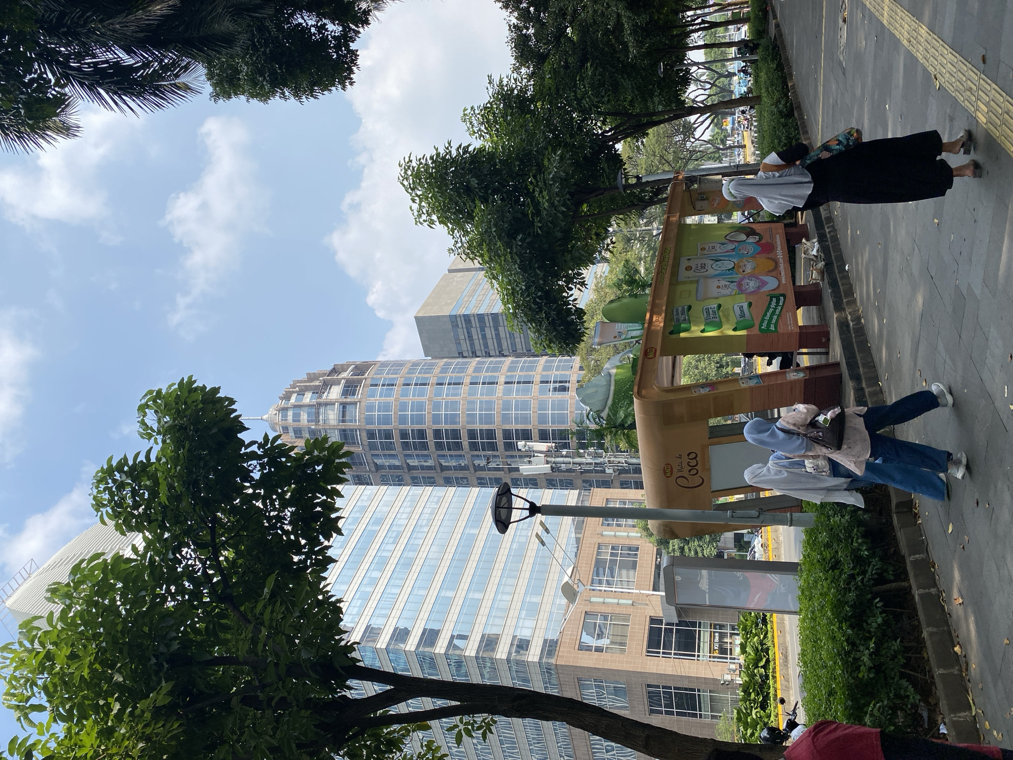 In South Jakarta, Indonesia, a street scene unfolds with a clear blue sky and scattered clouds overhead. Tall modern buildings with glass facades dominate the background, interspersed with trees that cast shadows on the sidewalk. In the foreground, a bus stop shelter with a distinctive curved roof displays advertisements, including one for "Nata de Coco." Two women wearing hijabs are walking on the sidewalk, one in a light-colored hijab and blue jeans, carrying a brown bag, and the other in a light blue hijab and a pink patterned top, also carrying a bag. Another woman, in a dark long dress and a light grey hijab, walks away from the camera. A person in a red shirt is partially visible on the far left. The sidewalk is paved with concrete and features a tactile paving strip. A street lamp stands near the bus stop. Lush green bushes line the edge of the sidewalk, and beyond them, a road with traffic is visible. The environment appears to be a well-maintained urban area.